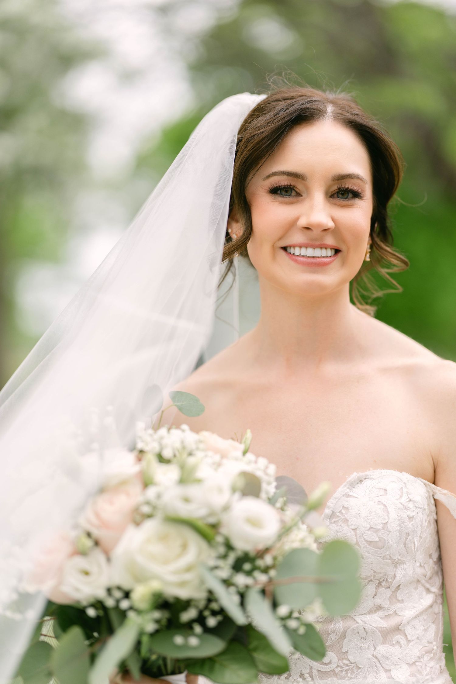 Smiling bride in strapless lace gown holding white floral bouquet with greenery, wearing a flowing veil outdoors.