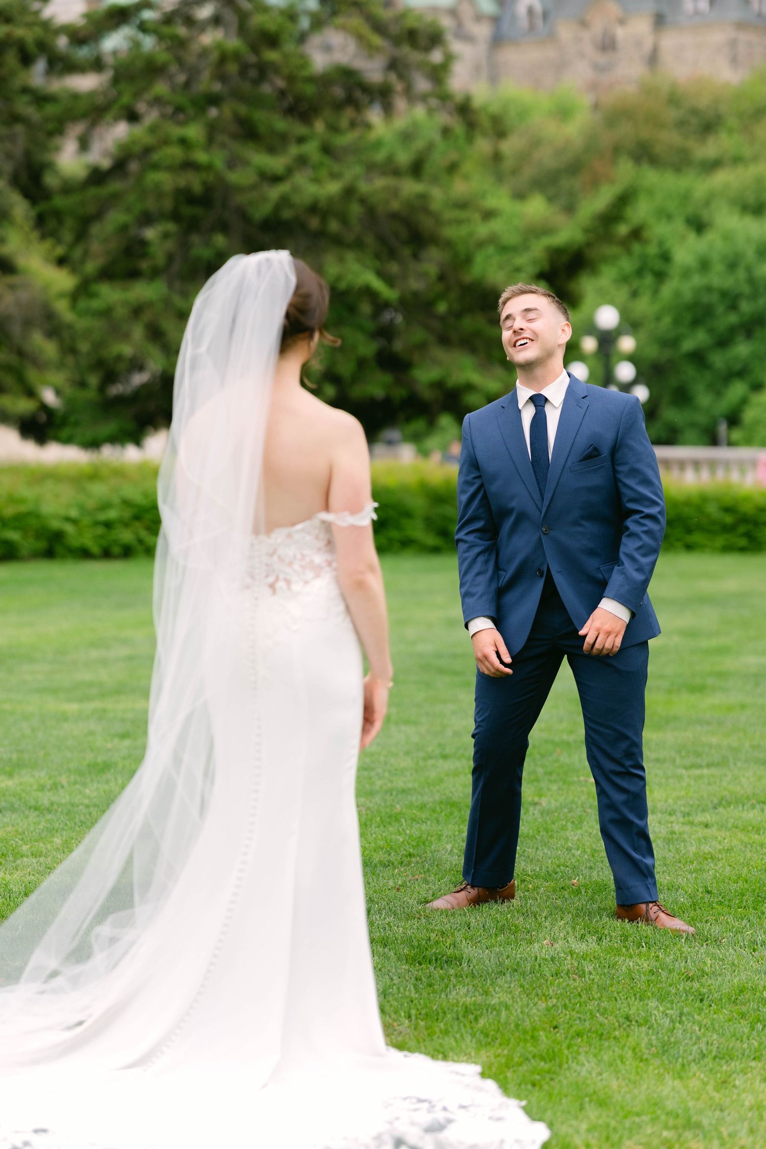 Groom in navy suit smiling joyfully at bride in lace wedding gown with veil during first look on green lawn.