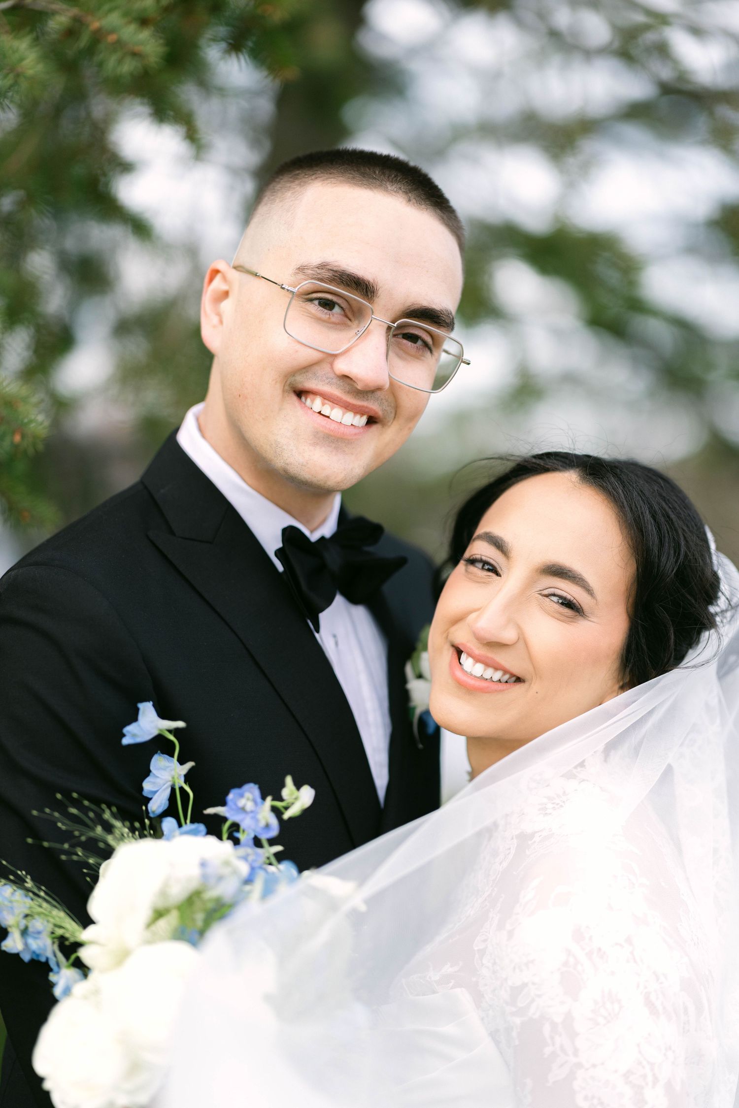 Smiling bride and groom posing together outdoors, groom wearing glasses and black bow tie, bride holding blue and white bouquet.