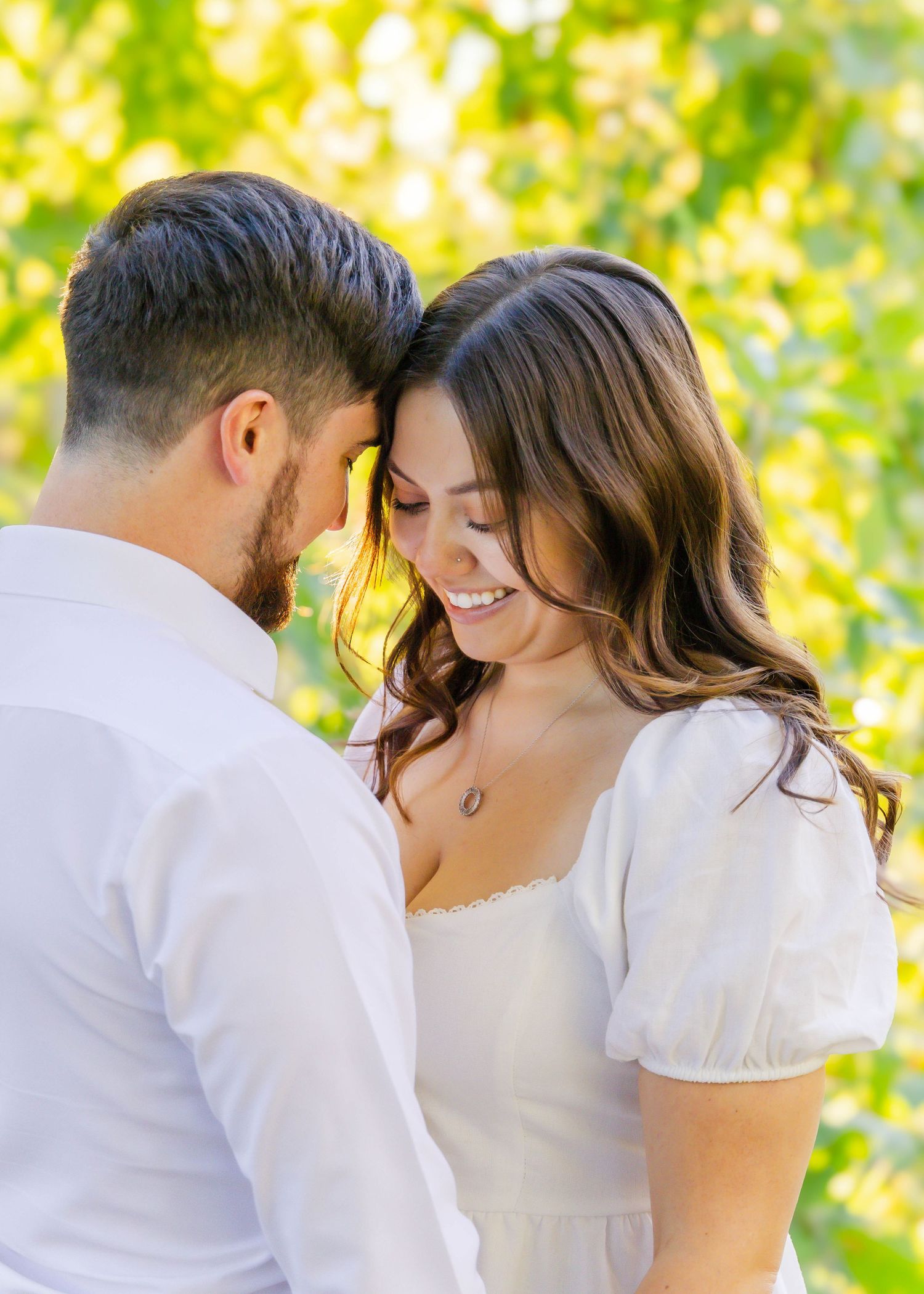 Couple in coordinated white outfits embrace in front of sunlit foliage during outdoor photo shoot.