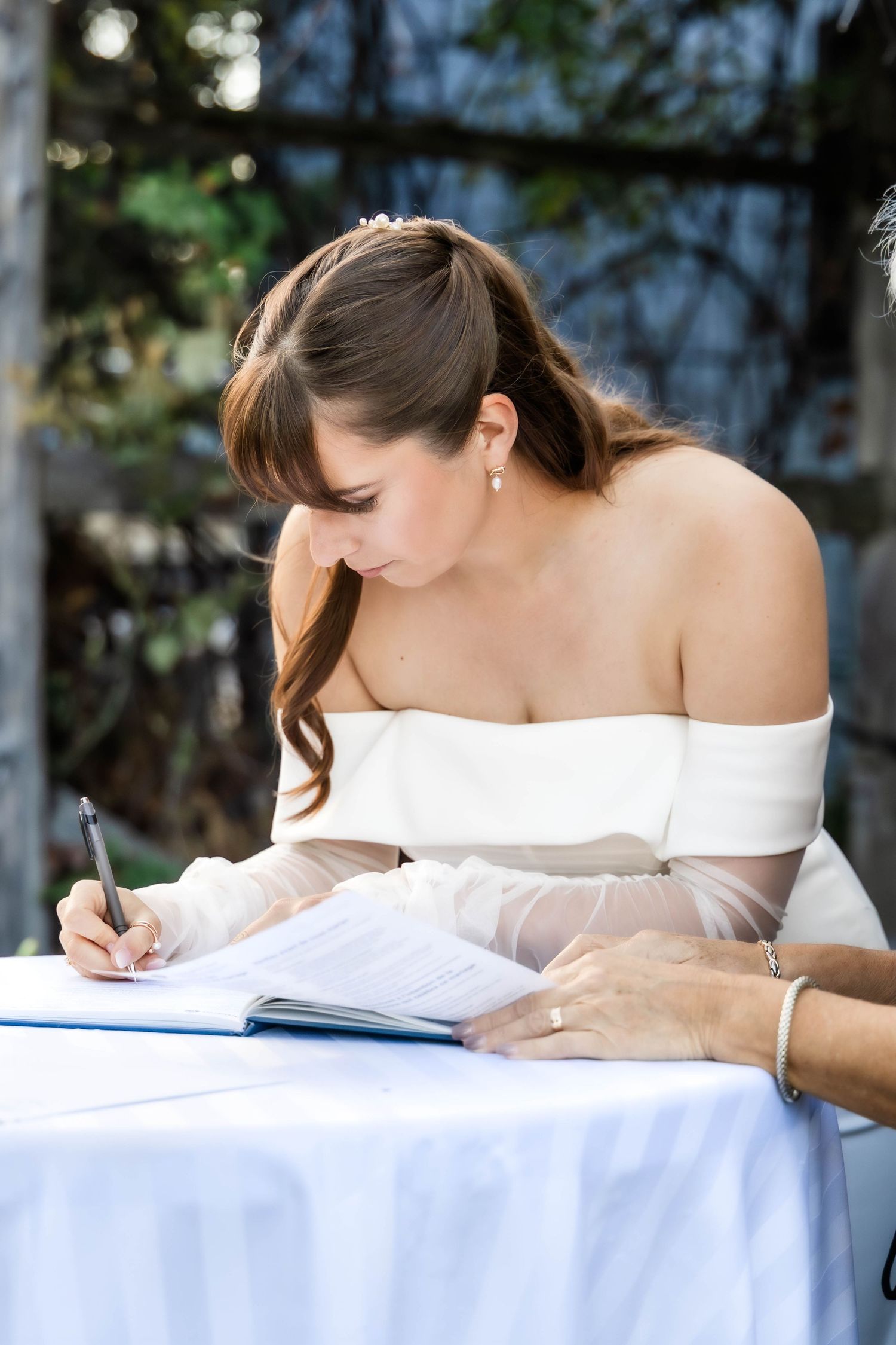 Bride wearing a white off-shoulder gown signs marriage documents at an outdoor table.