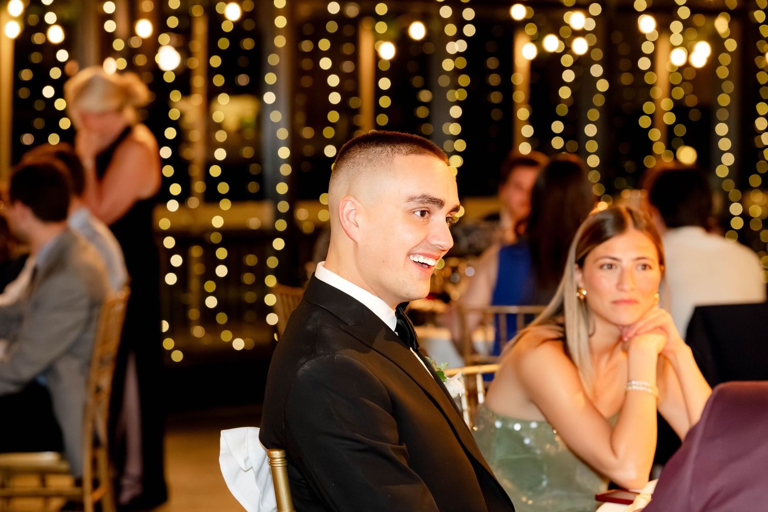 Man in tuxedo smiling at elegant event with bokeh string lights backdrop and guests seated nearby.