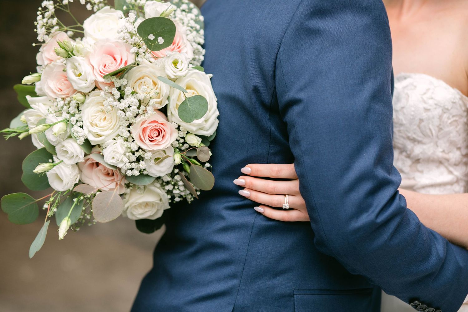 Bride holding a blush pink and white rose bouquet while embracing groom in a navy blue suit on wedding day.