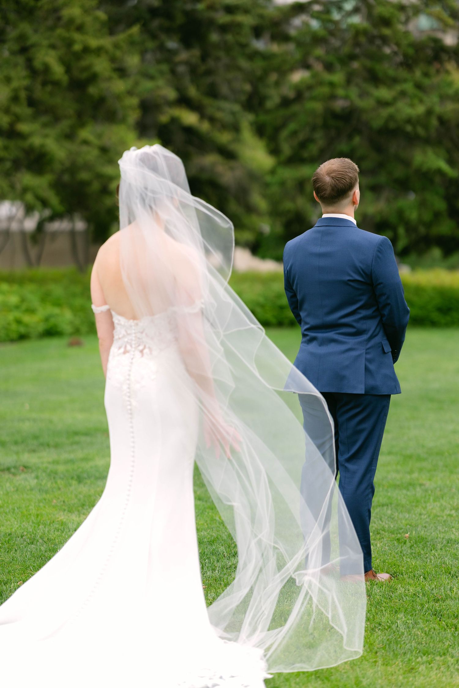 Bride in lace wedding gown with flowing veil walks beside groom in navy suit on green lawn.