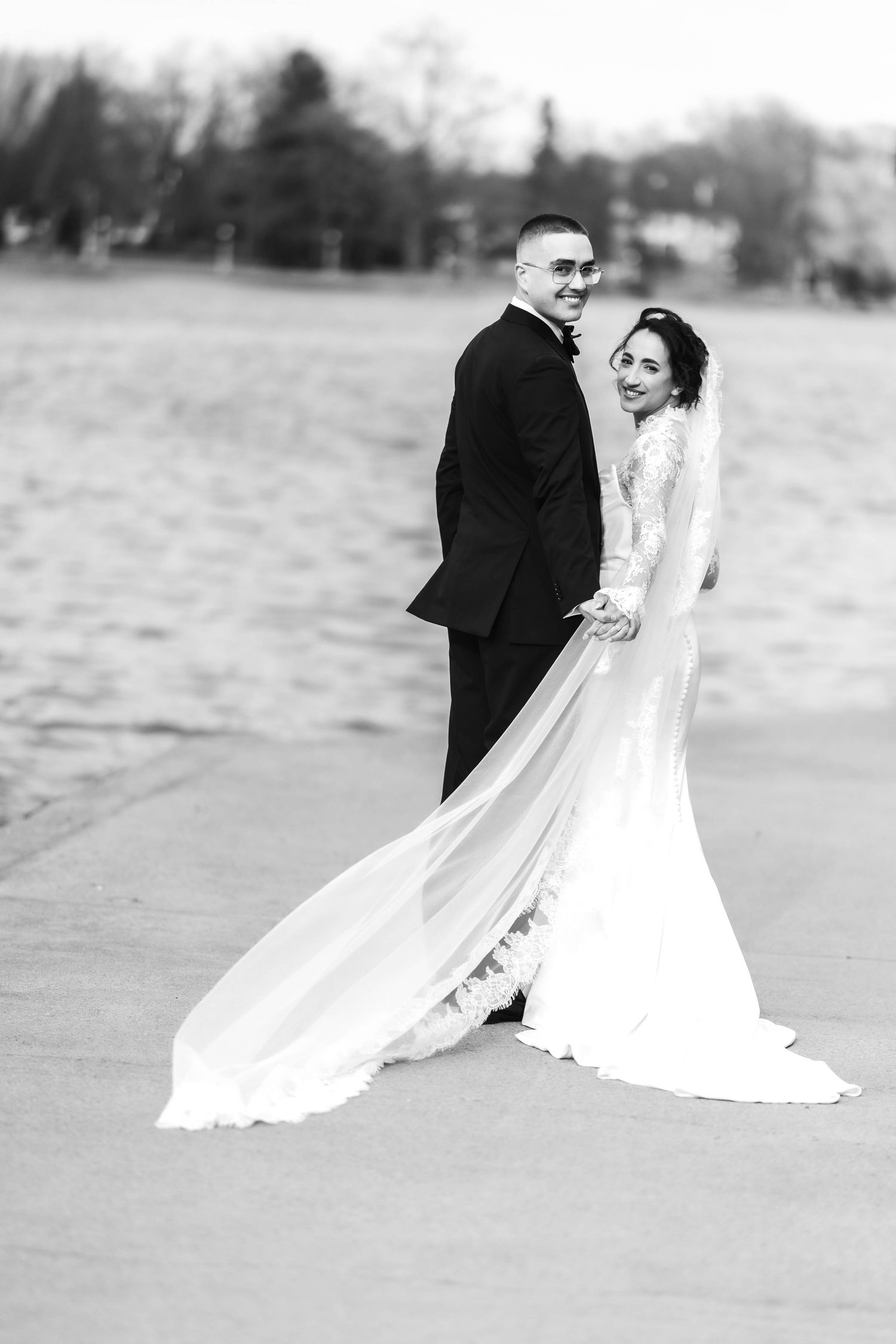 Black and white photo of a bride and groom holding hands near a lake, her veil flowing gracefully behind them.