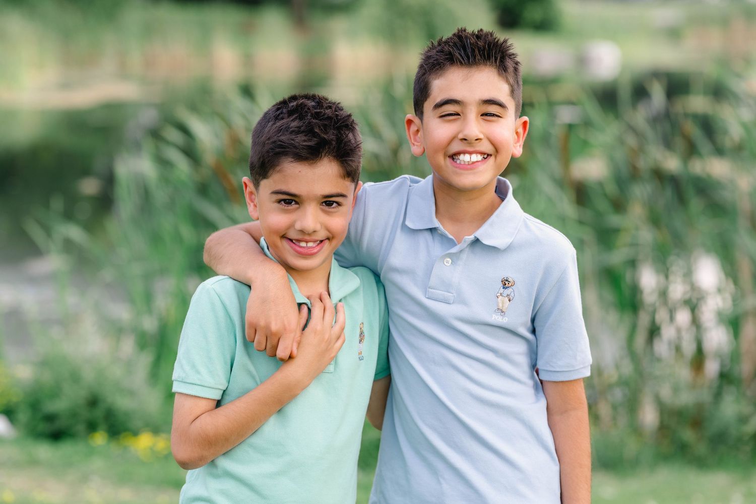 Two smiling boys in polo shirts embrace outdoors near a pond with green foliage in the background.