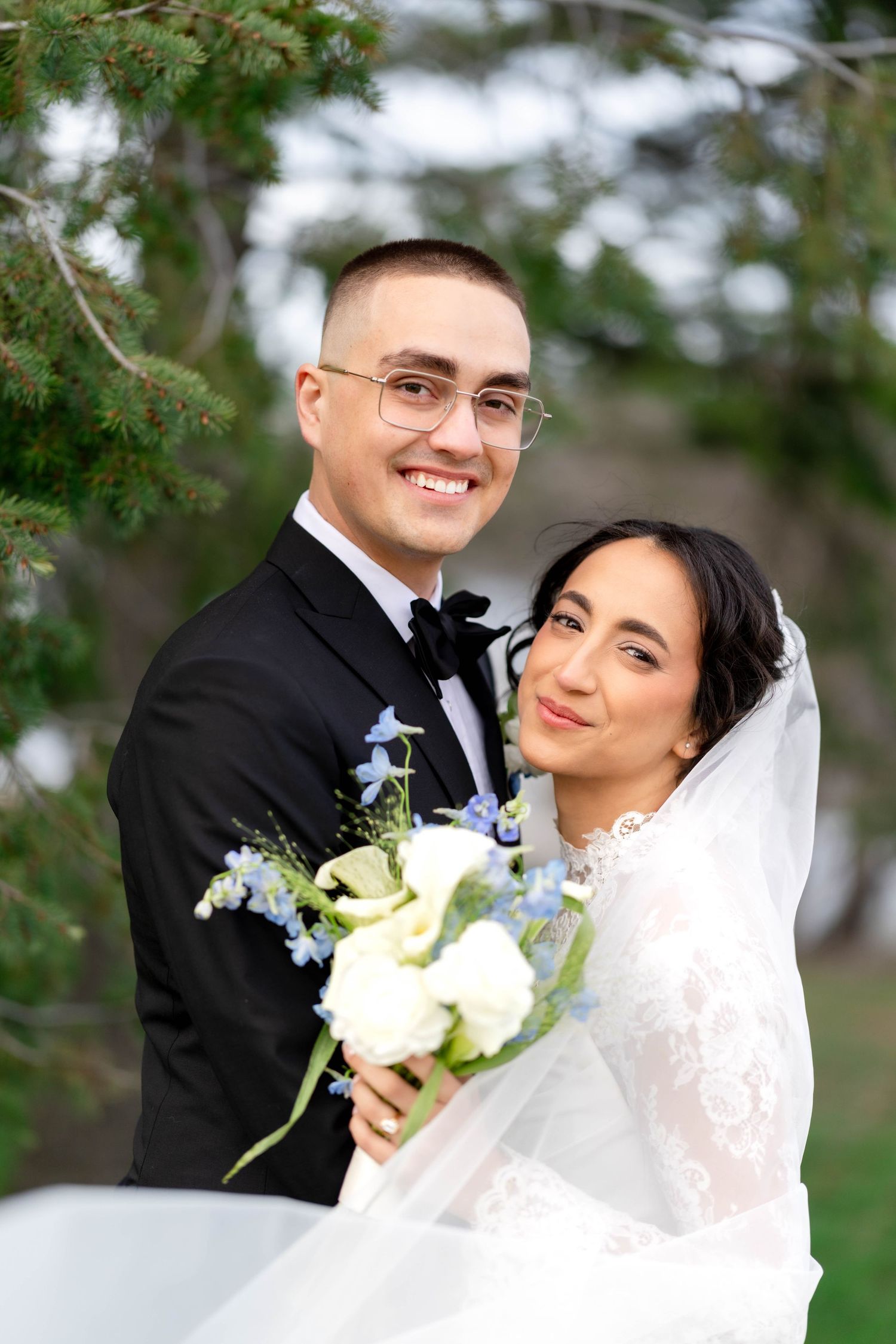 Smiling bride and groom pose outdoors, bride holding white and blue floral bouquet, groom in black tuxedo.