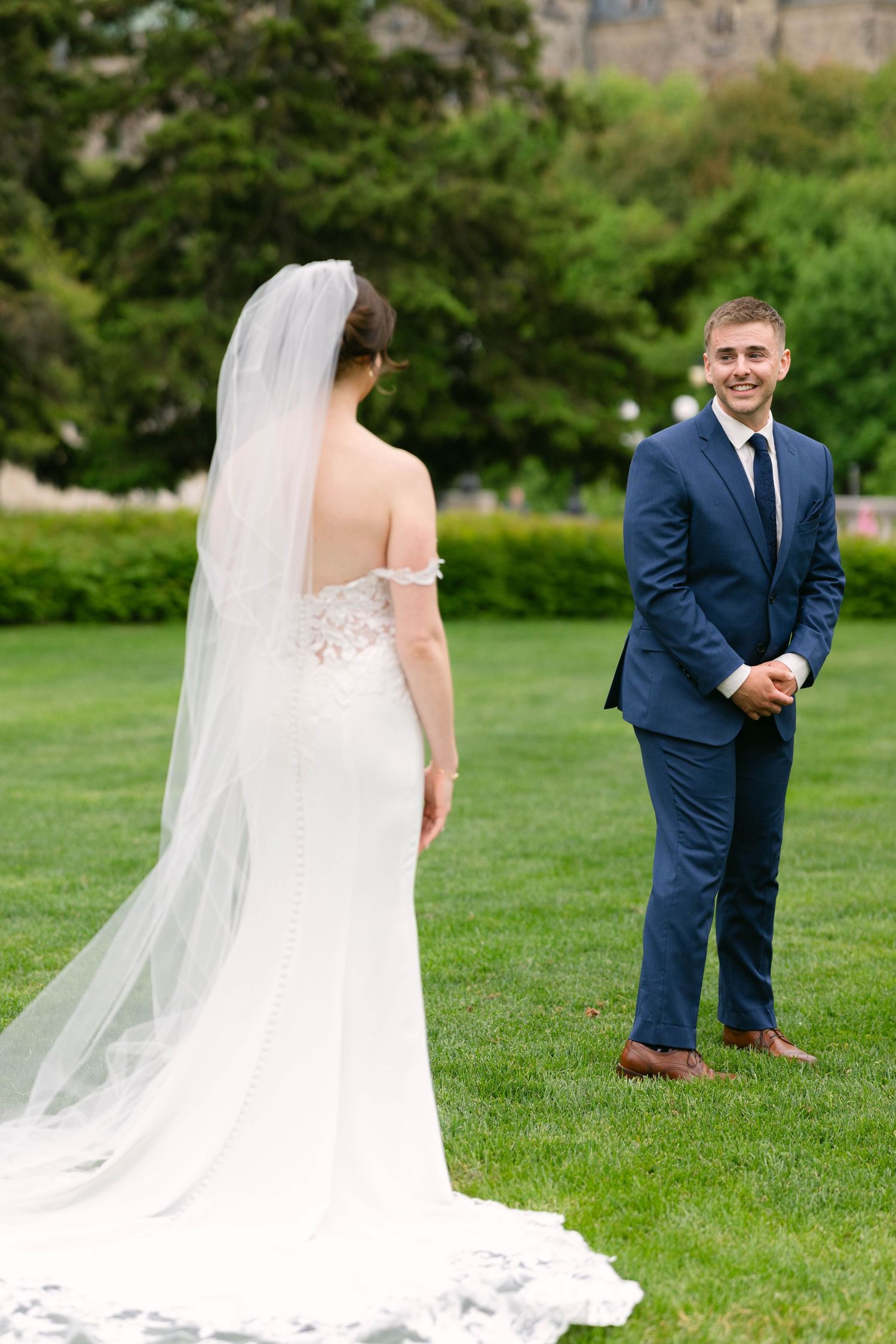 Bride in white gown and veil faces groom in navy suit during first look on green lawn.