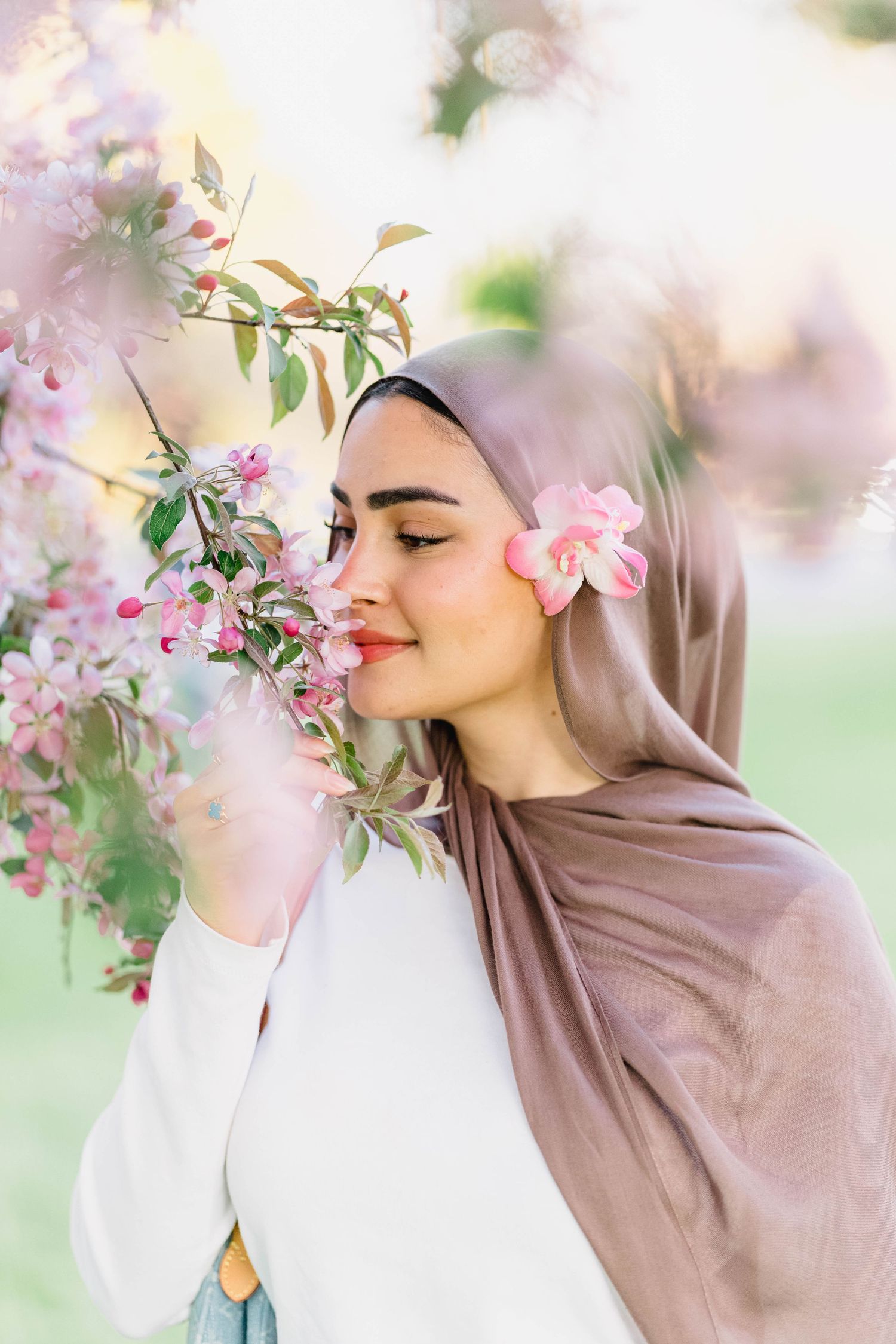 Young Muslim woman in hijab smelling pink cherry blossoms outdoors, with a flower tucked behind her ear.