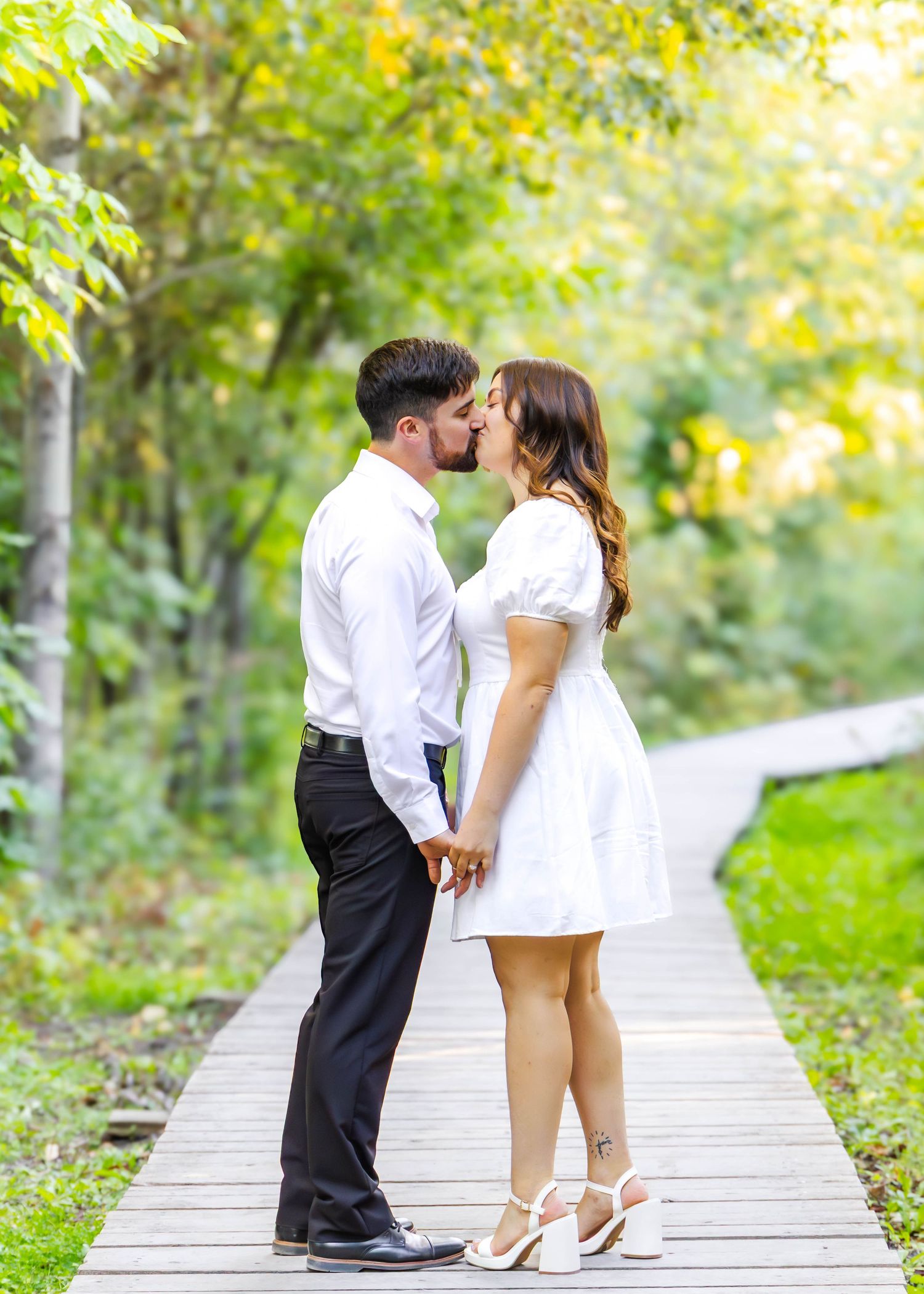 Couple in matching white outfits share sweet moments on wooden boardwalk surrounded by greenery.