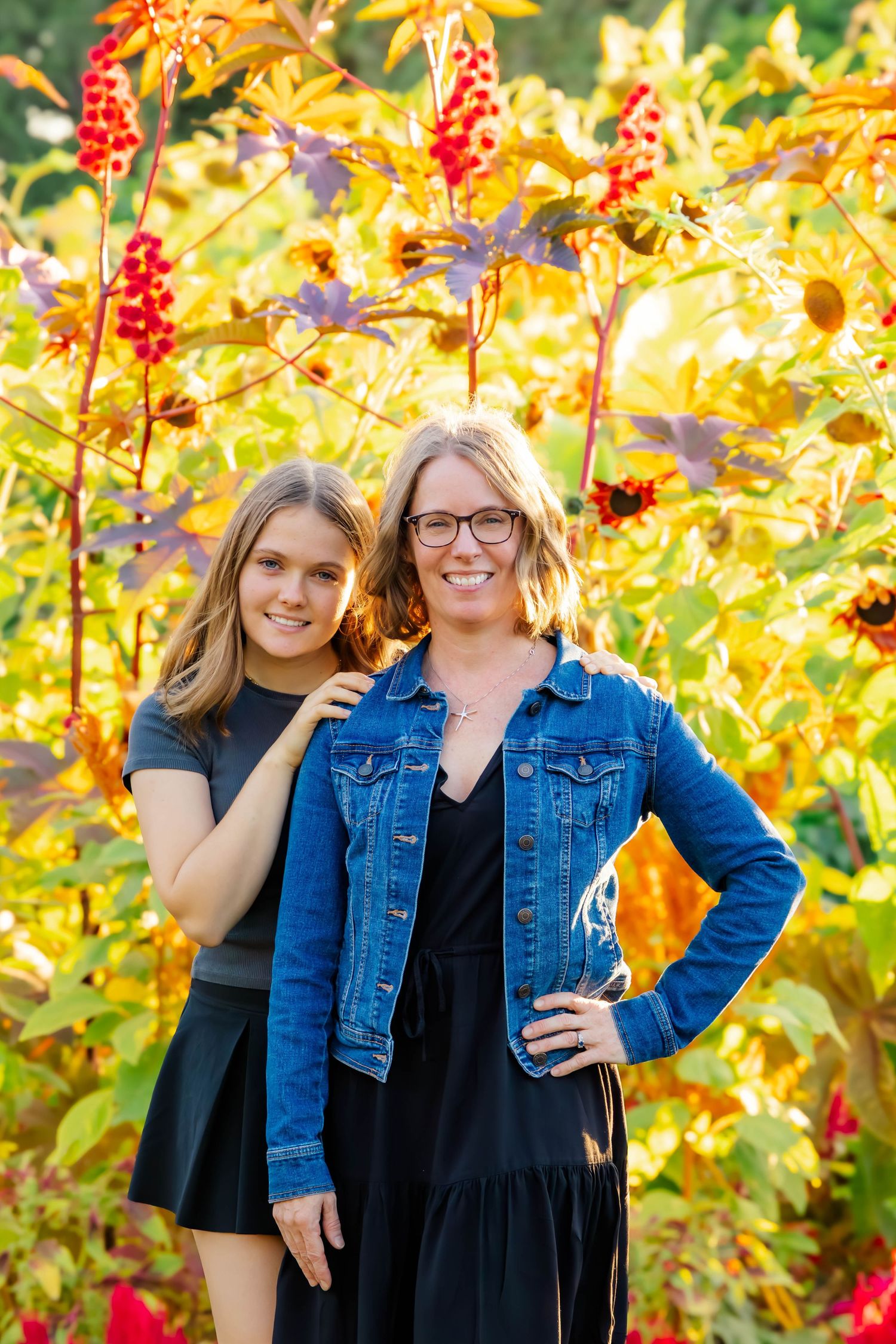 People wearing matching black dresses and denim jackets pose playfully in front of colorful autumn flowers.