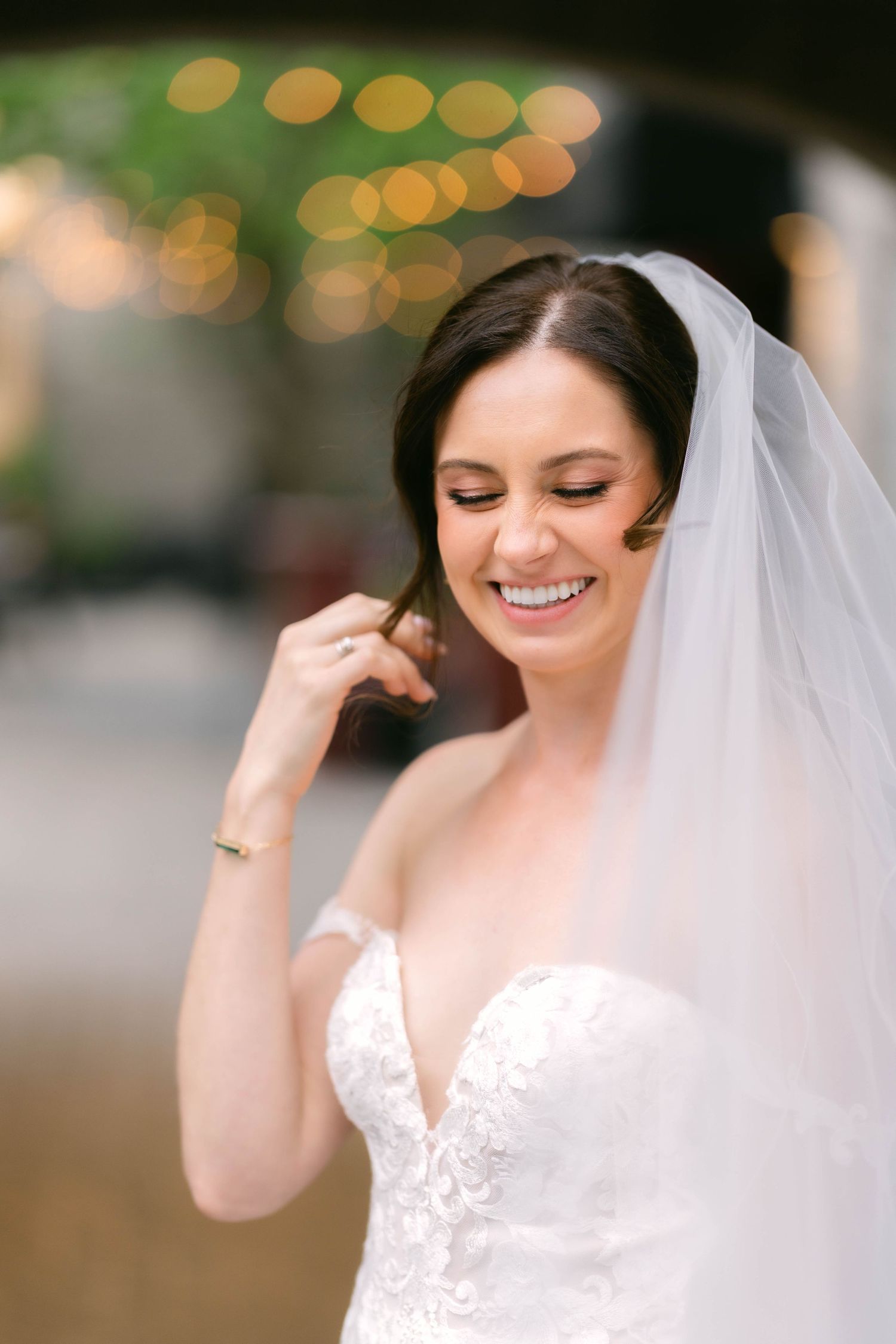 Radiant bride in lace strapless gown laughs softly while adjusting her veil with bokeh lights in background.