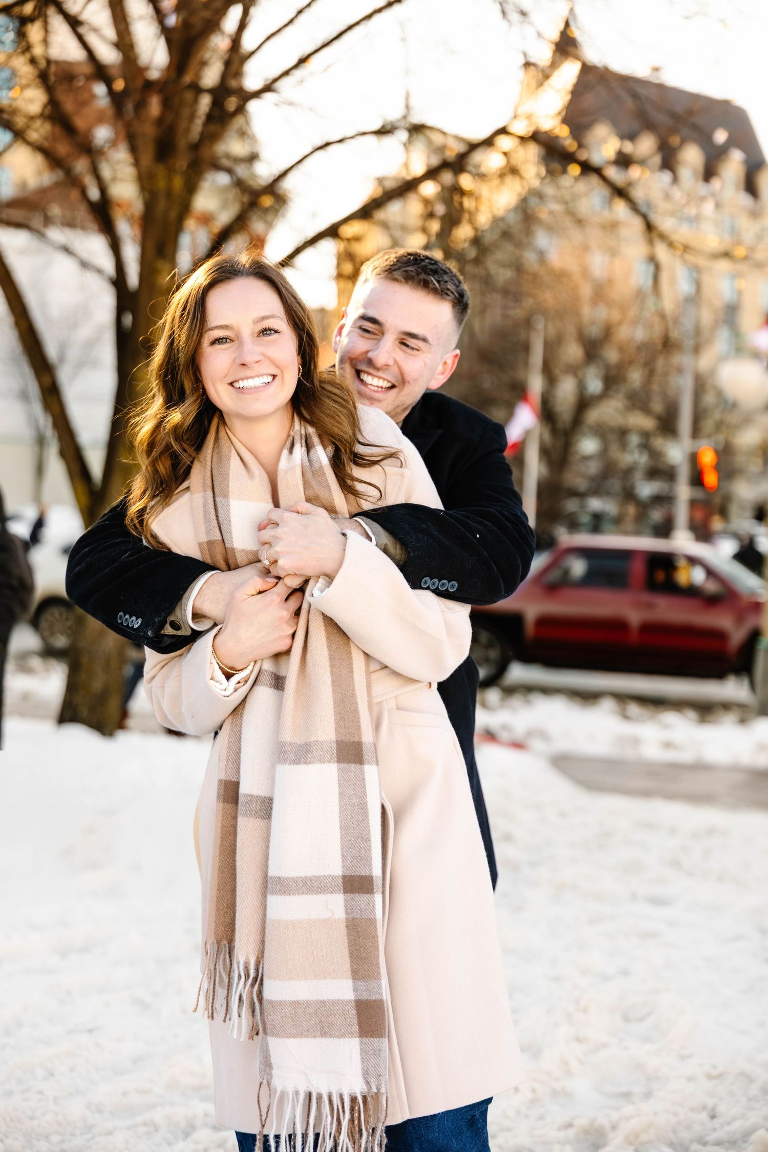 A couple shares a joyful embrace outdoors in winter, with snow on the ground and trees in the background.