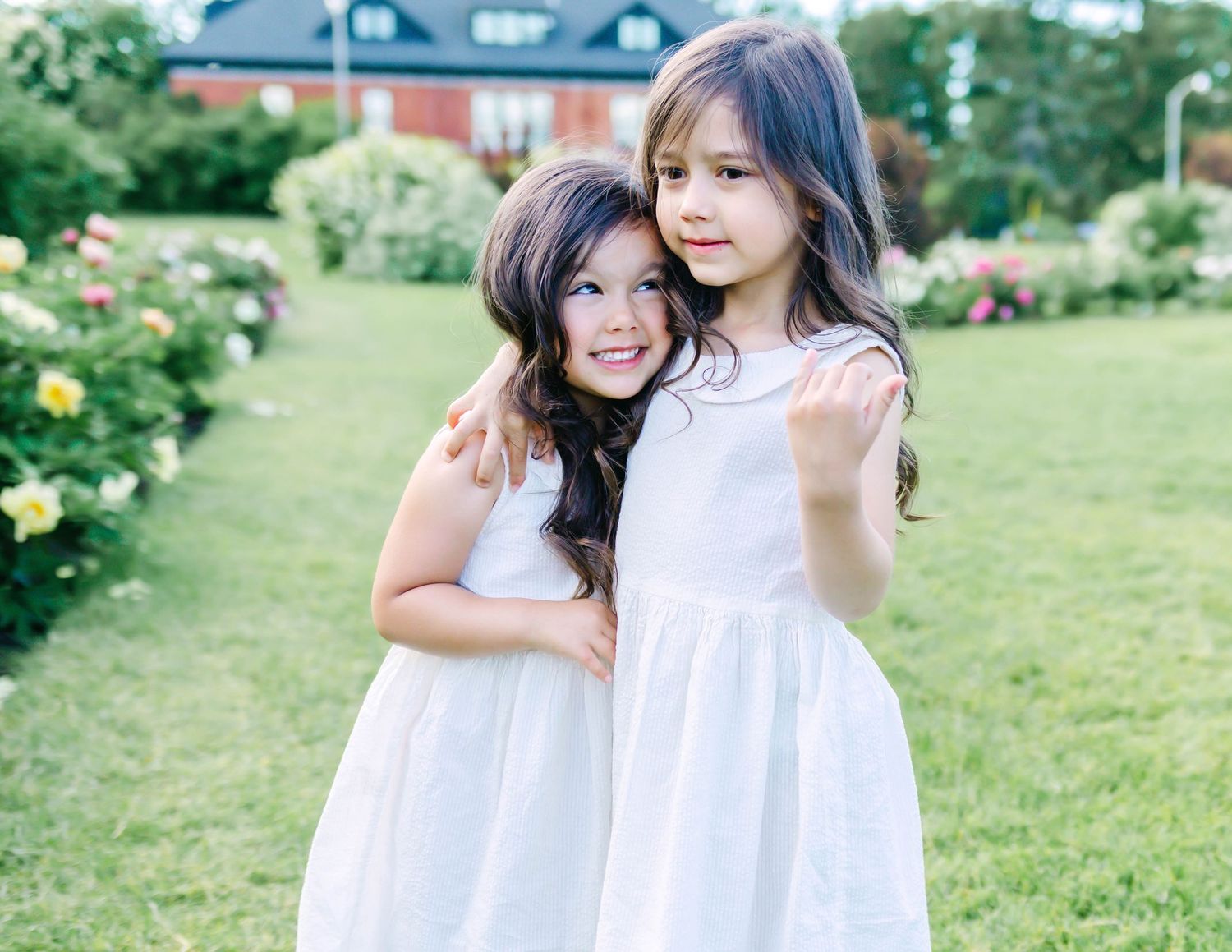 Two people in white dresses embrace while standing in a sunny garden with blooming flowers and green grass.