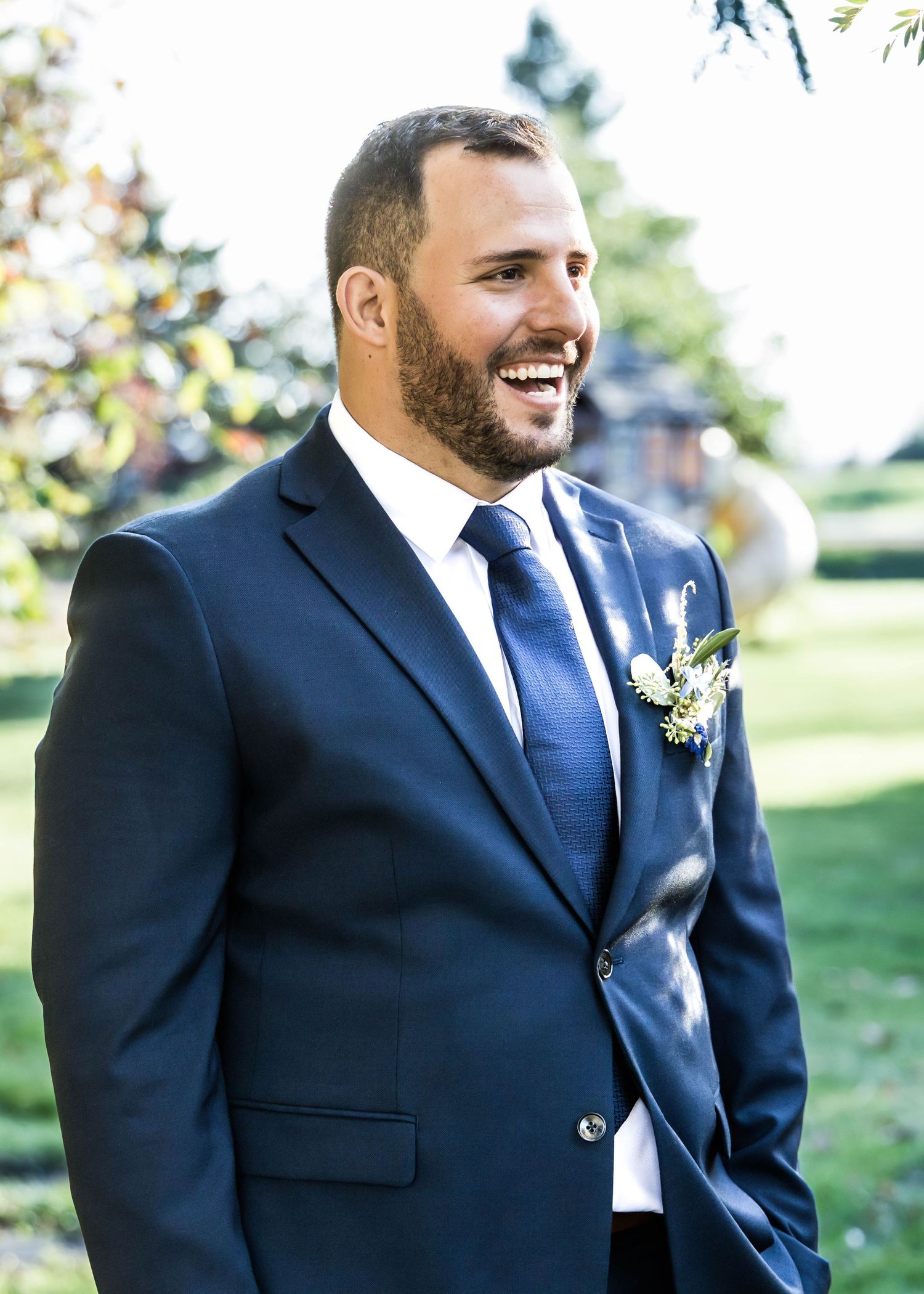 Groom in navy blue suit and white tie smiles during outdoor wedding photo session in natural setting.