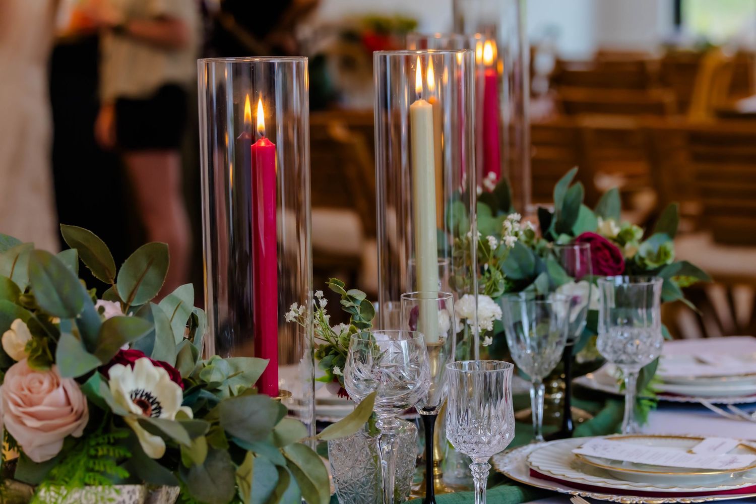 Elegant wedding reception table setting with crystal glasses, burgundy candles, and eucalyptus greenery centerpieces.