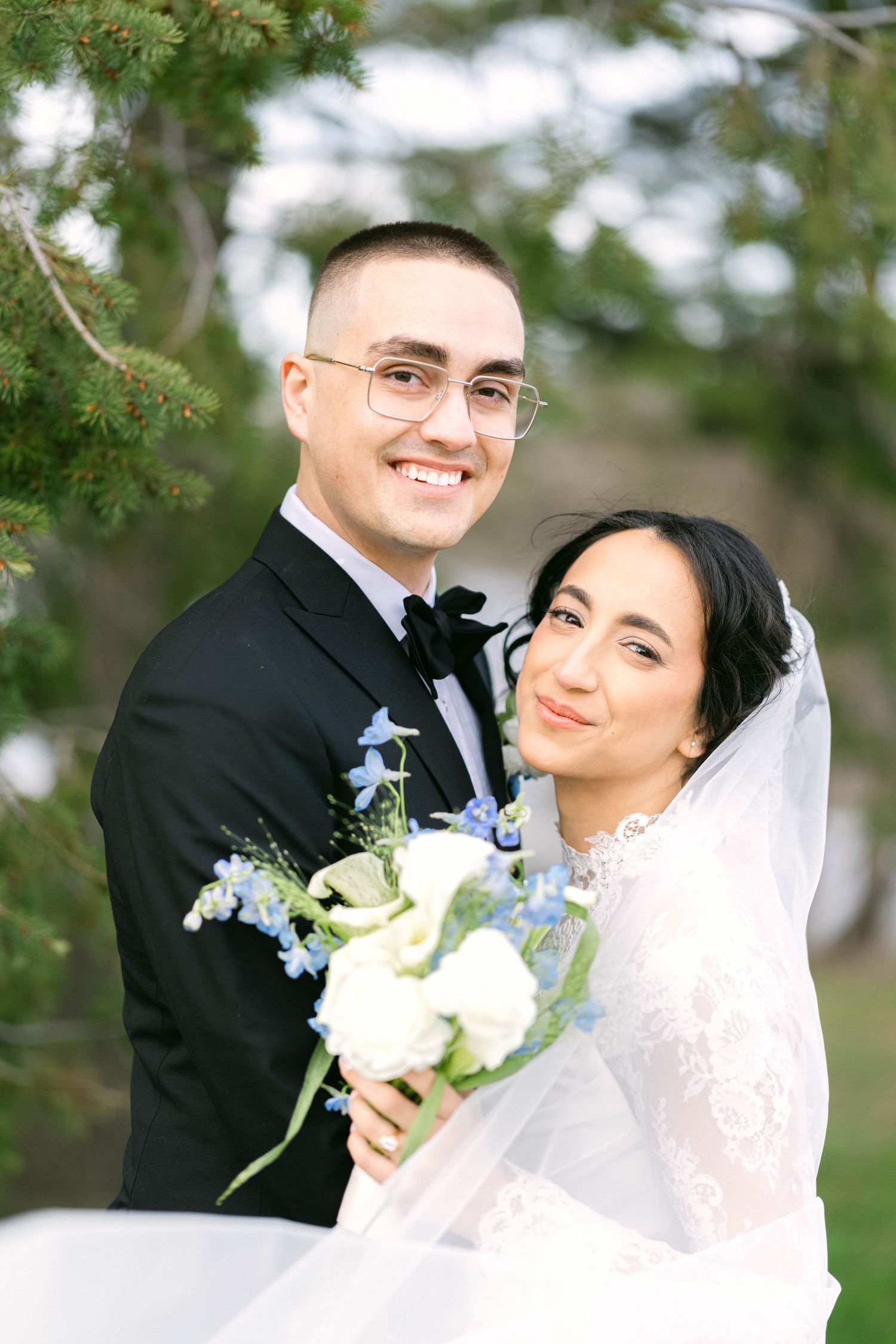 Smiling bride and groom pose together outdoors, she holds white and blue bouquet while her veil flows gracefully.
