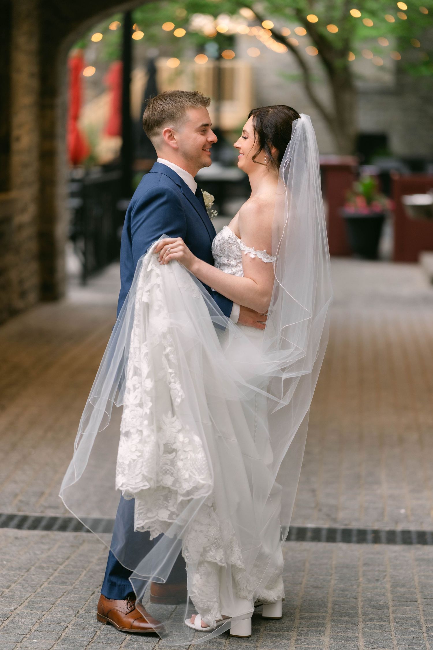 Groom lifts bride in a lace wedding dress and long veil under string lights in a charming outdoor venue.