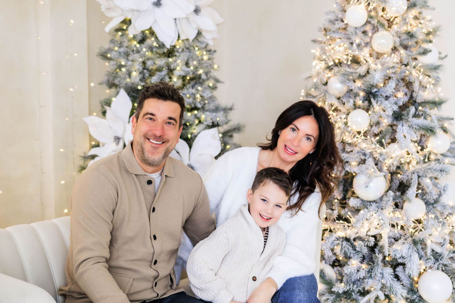 A family poses together on a white couch between two decorated Christmas trees with white ornaments and lights.