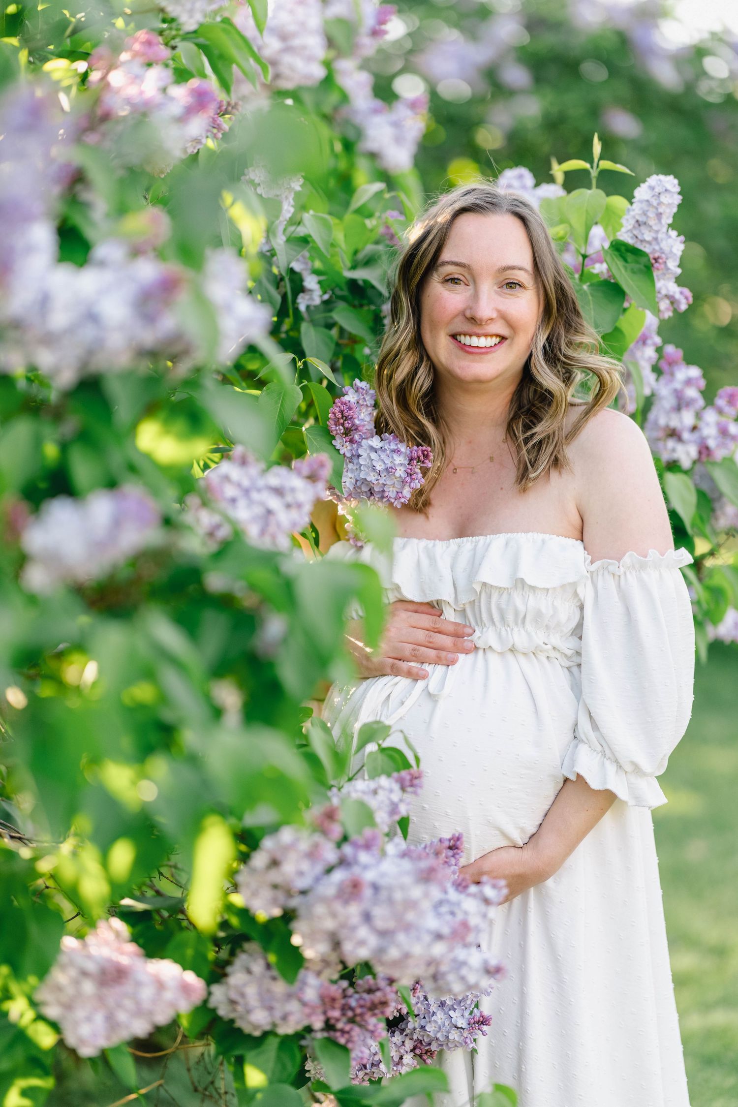 Smiling pregnant woman in white off-shoulder dress standing among blooming lilac bushes in a lush green garden.