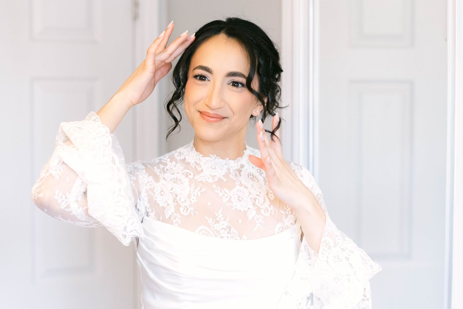 A bride in a white lace wedding dress smiles while adjusting her hair, posing elegantly in front of white doors.