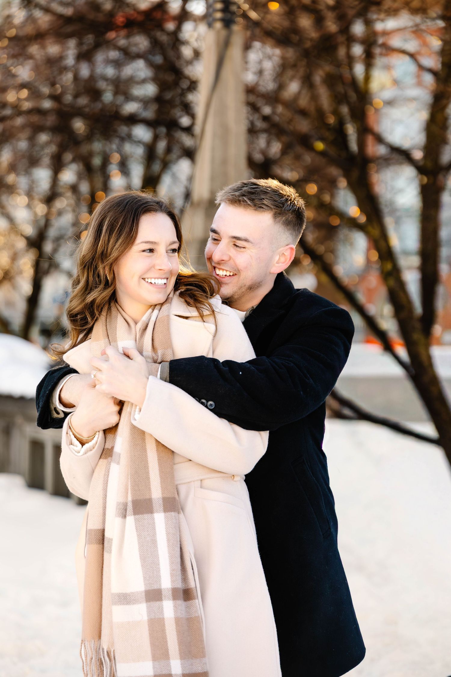 A couple shares a playful moment in the snow, wearing winter attire with a black coat and beige scarf.