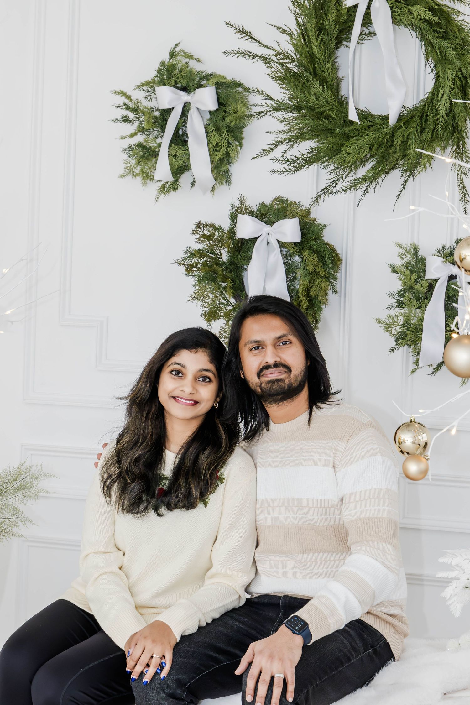 Couple in cream sweaters poses for holiday photos with evergreen wreaths and gold decorations.