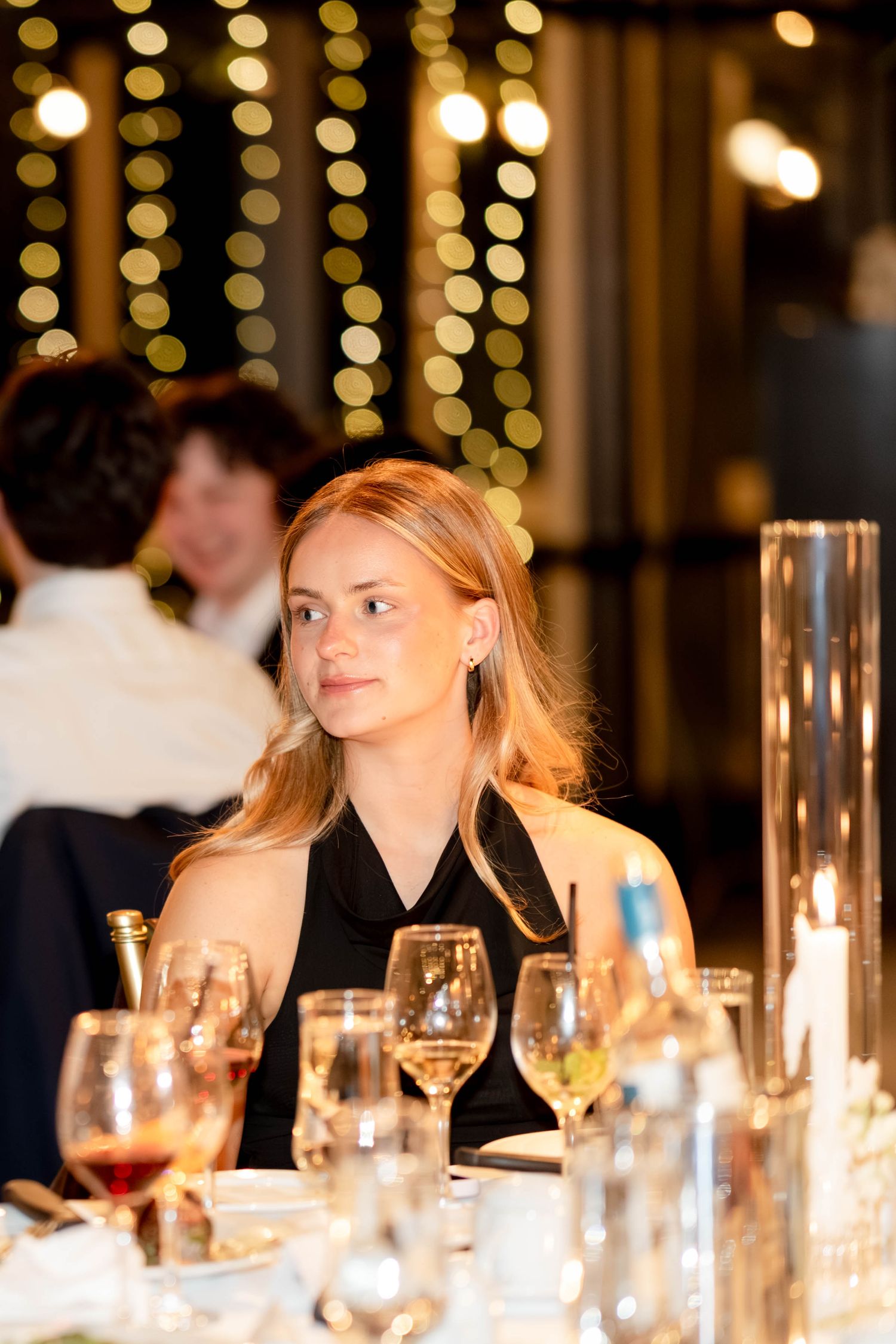 Elegant woman in black dress seated at a candlelit dinner table with wine glasses and bokeh lights in background.