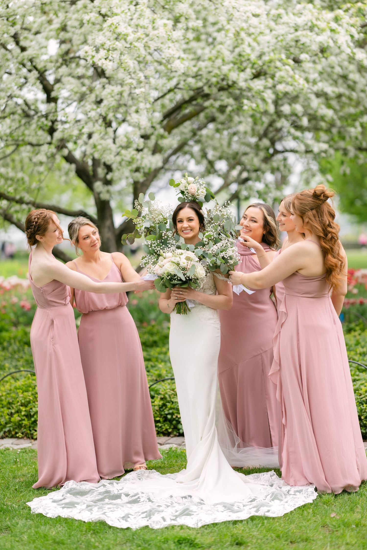 Bride in white gown holds bouquet surrounded by bridesmaids in pink dresses under blooming trees.