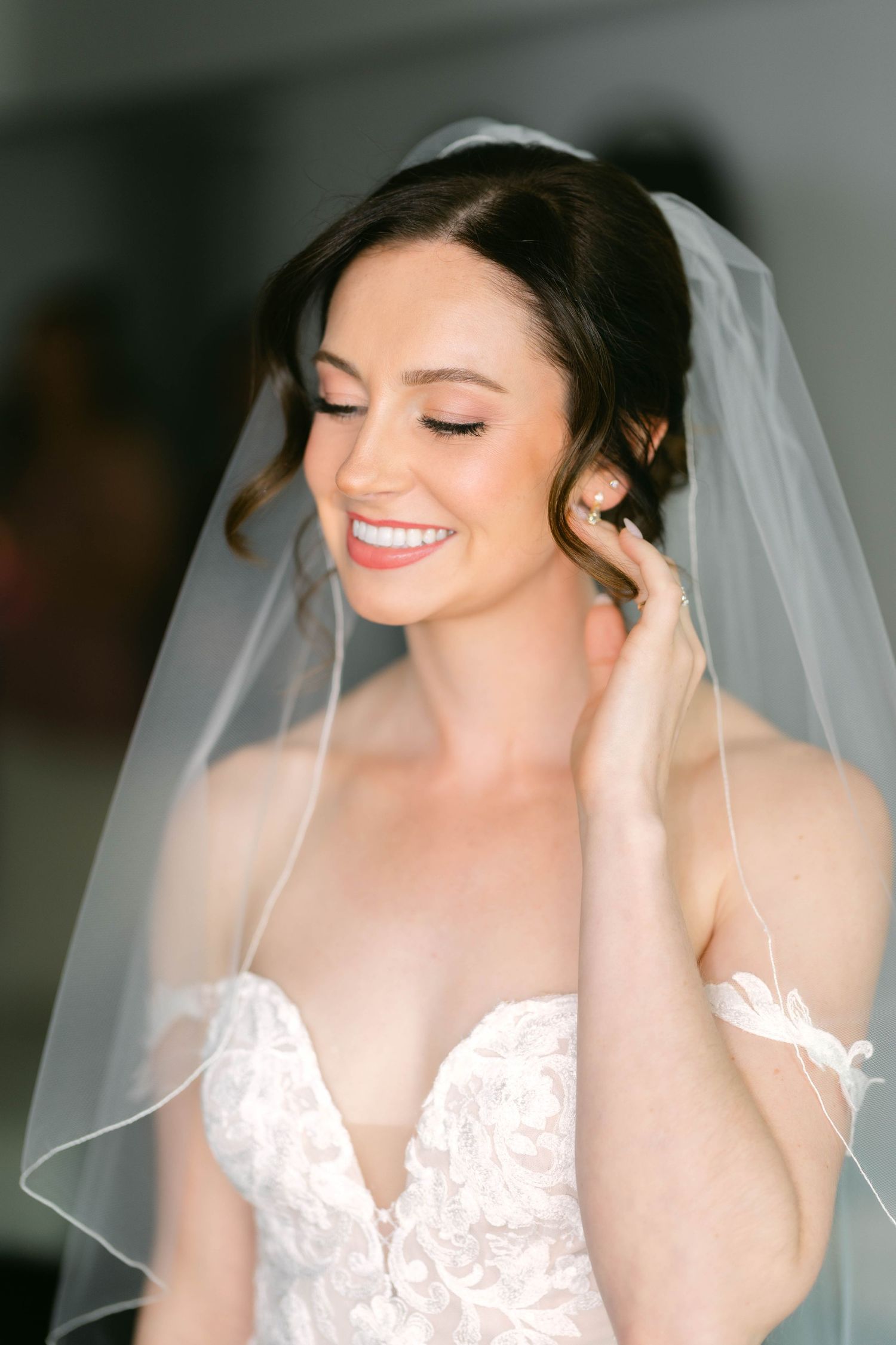 Smiling bride in lace gown and white veil touches her ear, showcasing elegant bridal makeup and updo hairstyle.