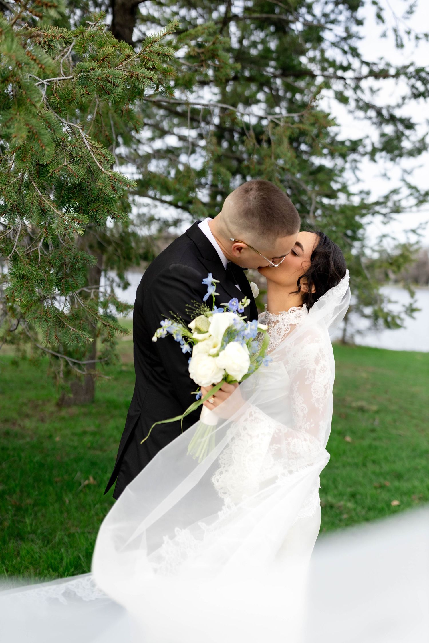 Bride and groom share romantic dip kiss outdoors, bride in lace dress with flowing veil holding white and blue bouquet.