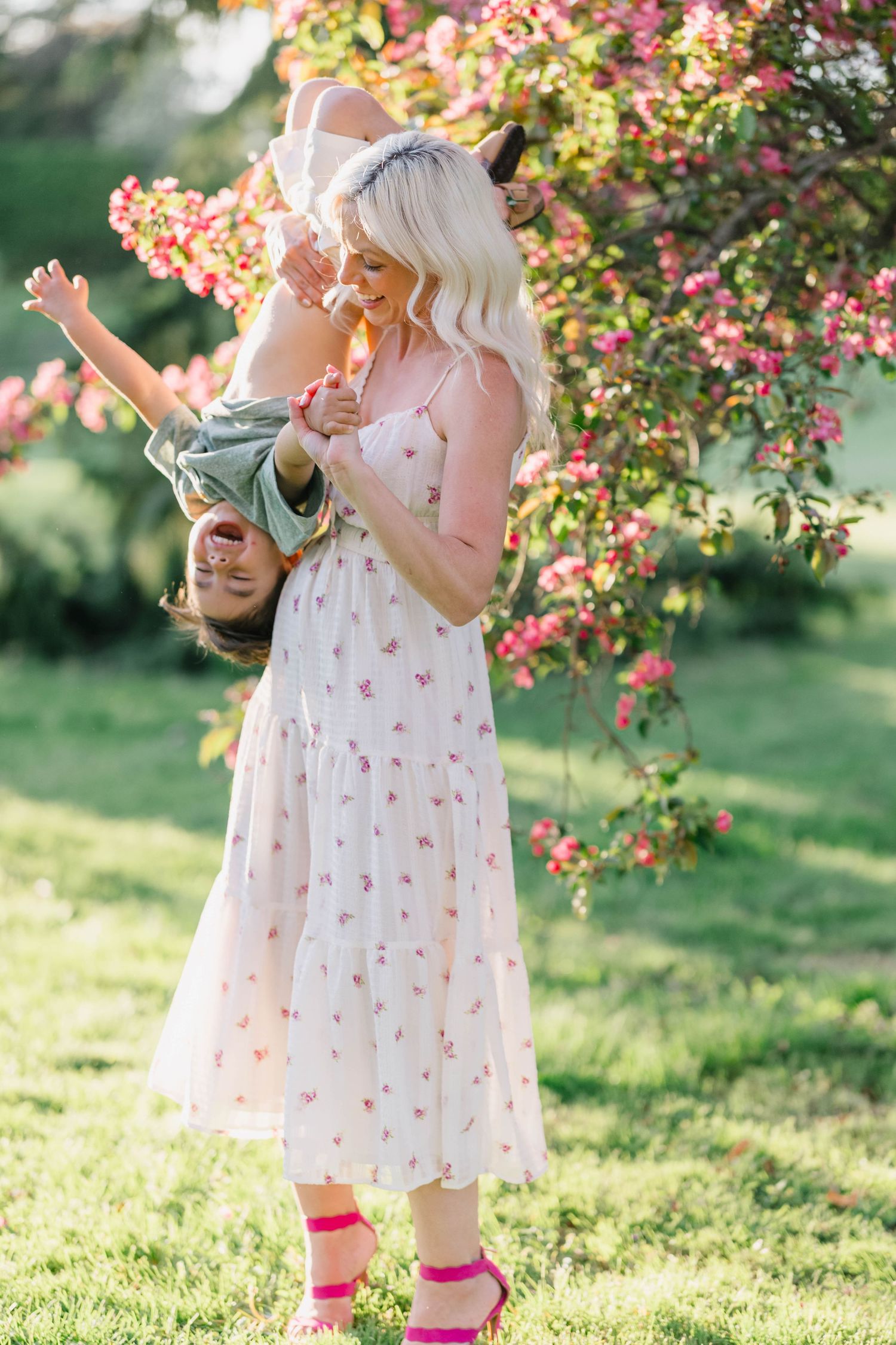 A blonde woman in a floral dress playfully holds a laughing child upside down near pink blooming trees outdoors.