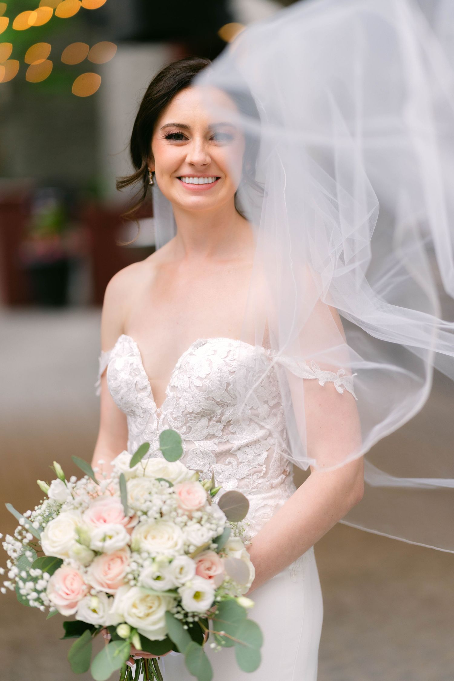 Bride smiling in lace strapless gown, holding blush and white rose bouquet with veil flowing around her.