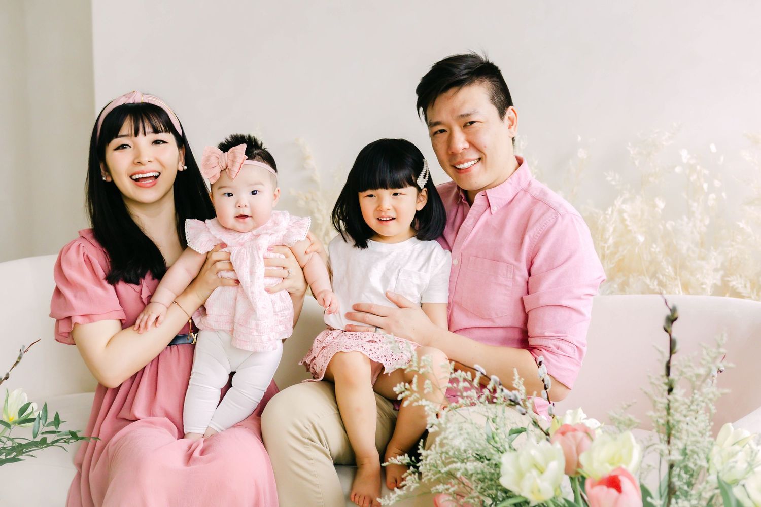 Family wearing coordinated pink and beige outfits sitting together in a bright studio setting with floral decorations.