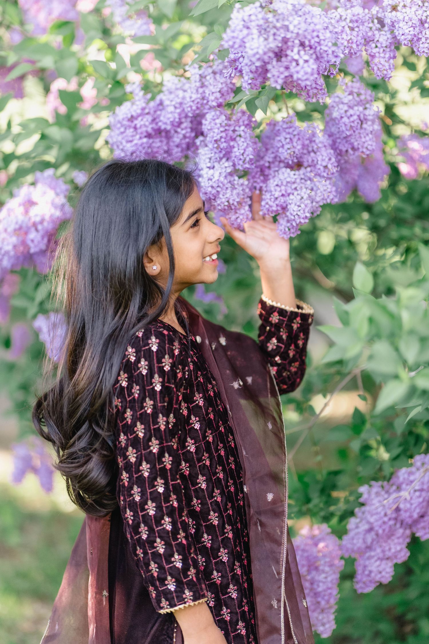 Young girl in dark floral kurta smiling among blooming purple lilac flowers in a lush garden.