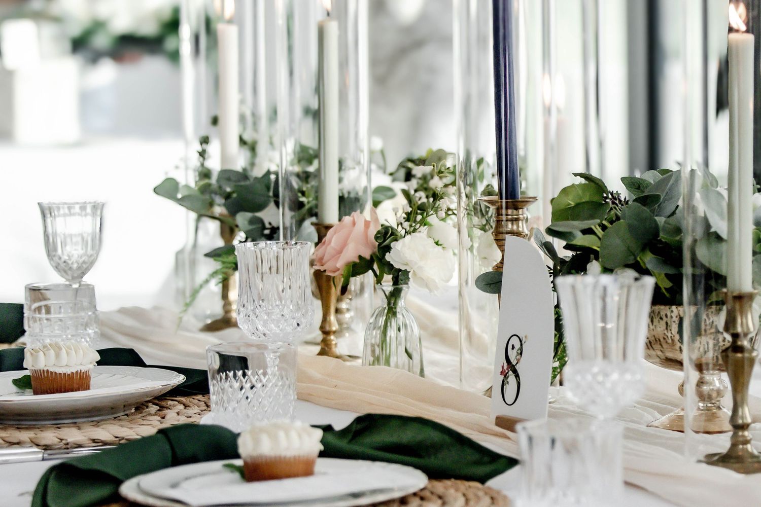 Elegant wedding reception table settings with white linens, crystal glassware, and emerald green accents in a bright venue.