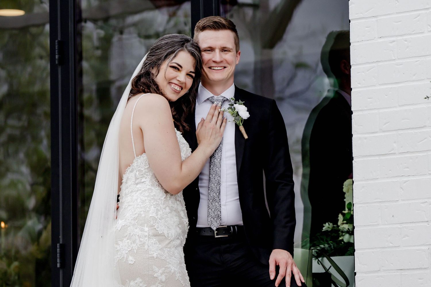 Wedding couple poses together against a white wall backdrop with elegant formal attire.