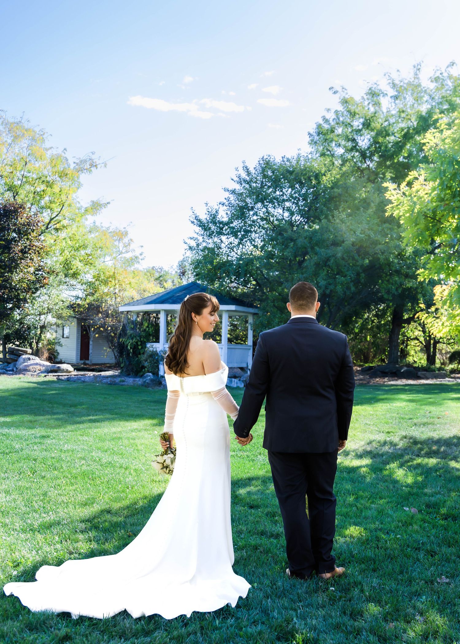 A couple in wedding attire holds hands while standing on a lush green lawn with trees and a gazebo in the background.