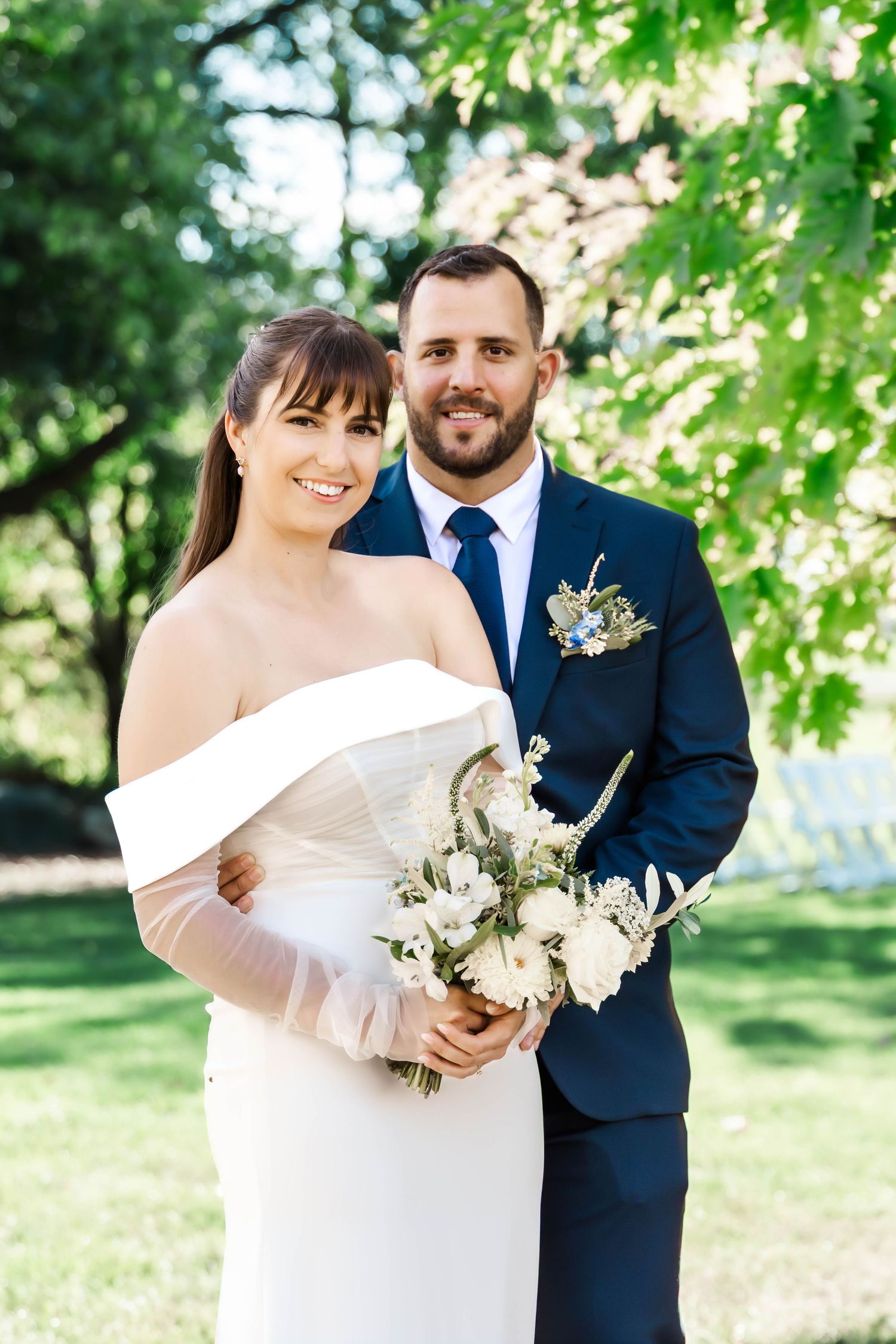 A couple in wedding attire embraces outdoors with lush greenery and natural lighting in a romantic garden setting.
