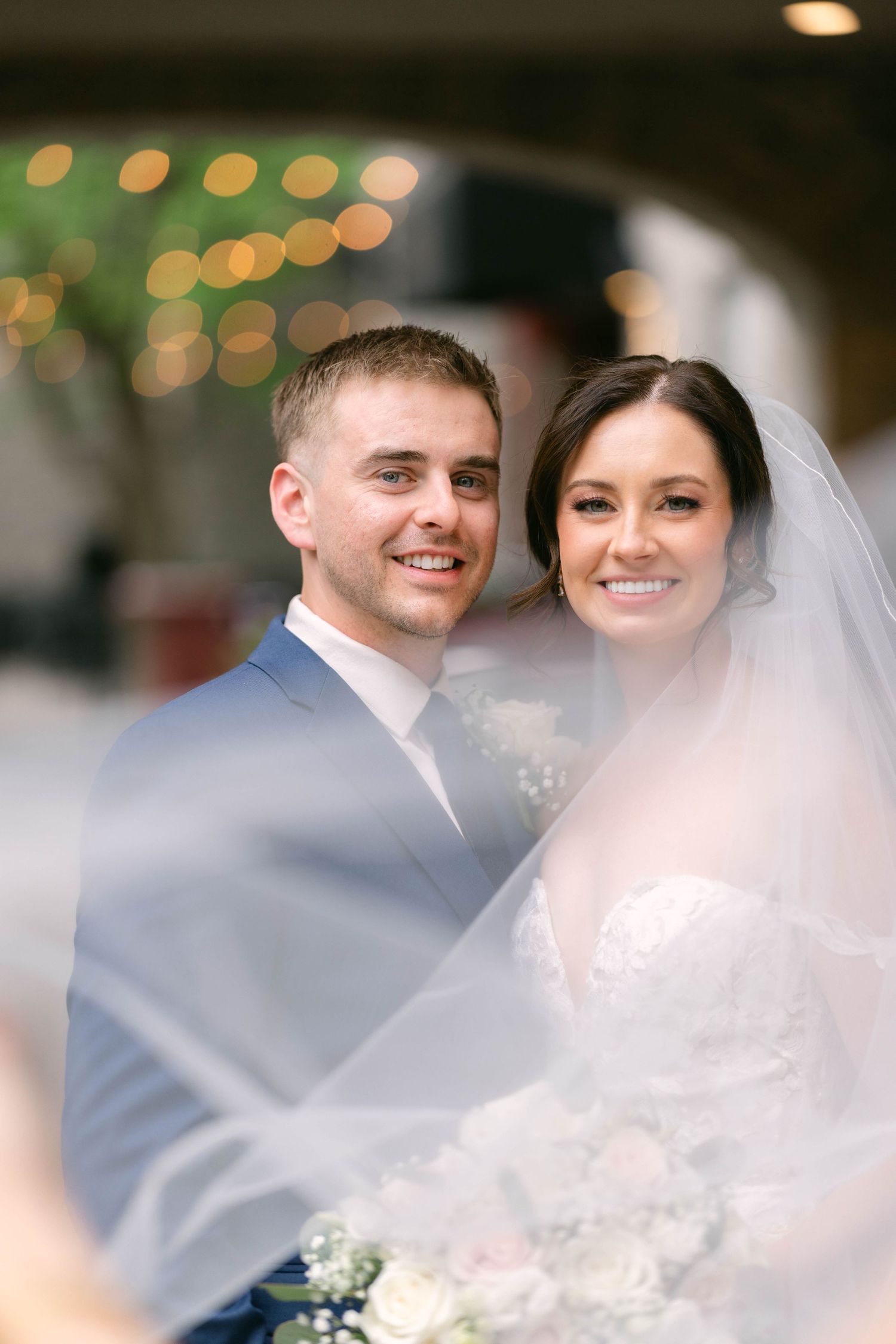 Smiling bride and groom posing together under a flowing veil, with bokeh lights in the background at their wedding.