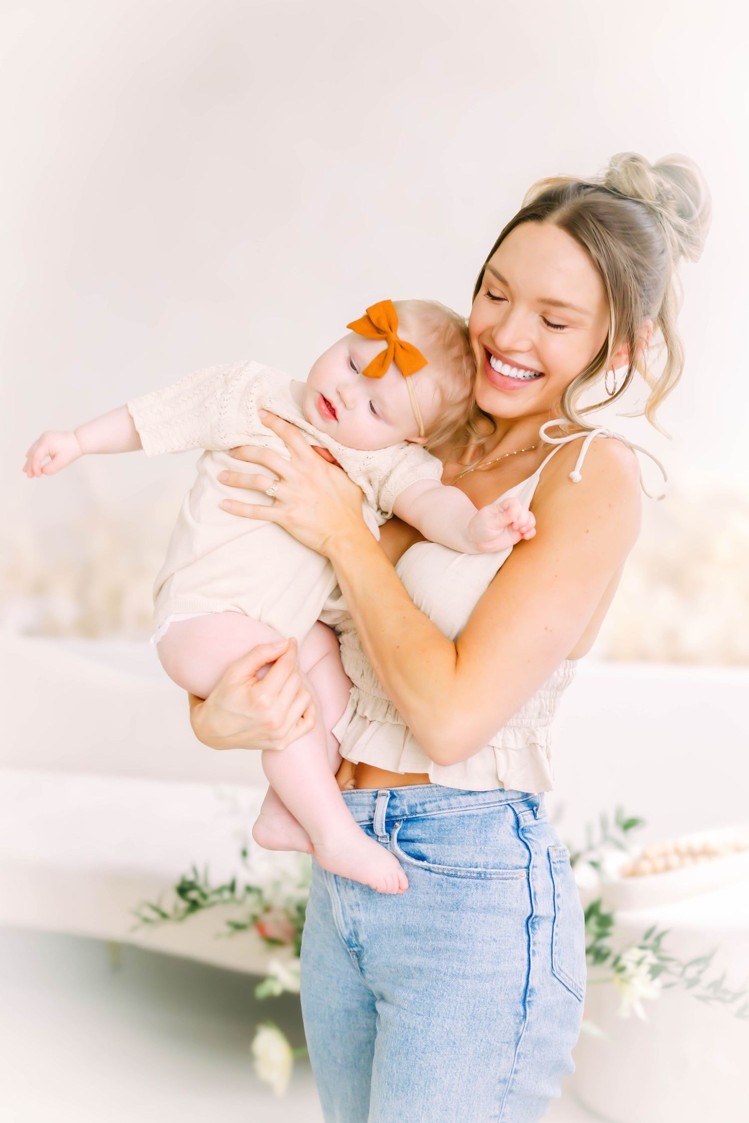 Mother in jeans and pink top holds baby in soft pink outfit in bright indoor setting with three sequential poses.