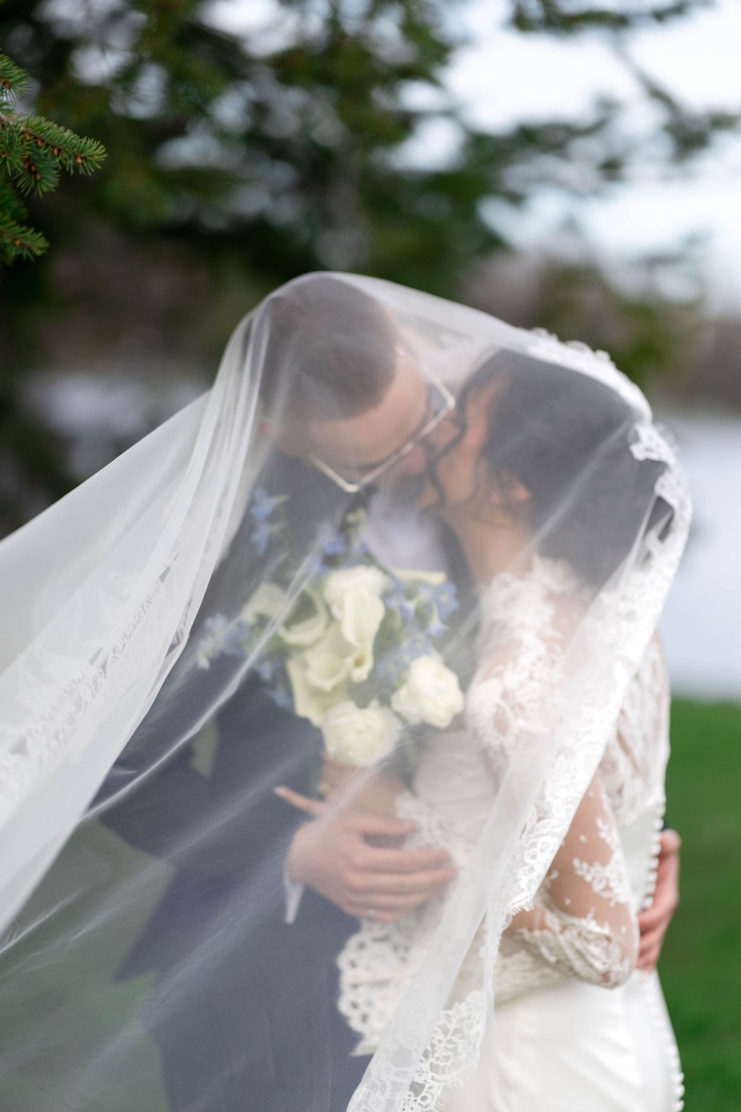 Bride and groom sharing a romantic kiss under a flowing white wedding veil outdoors.