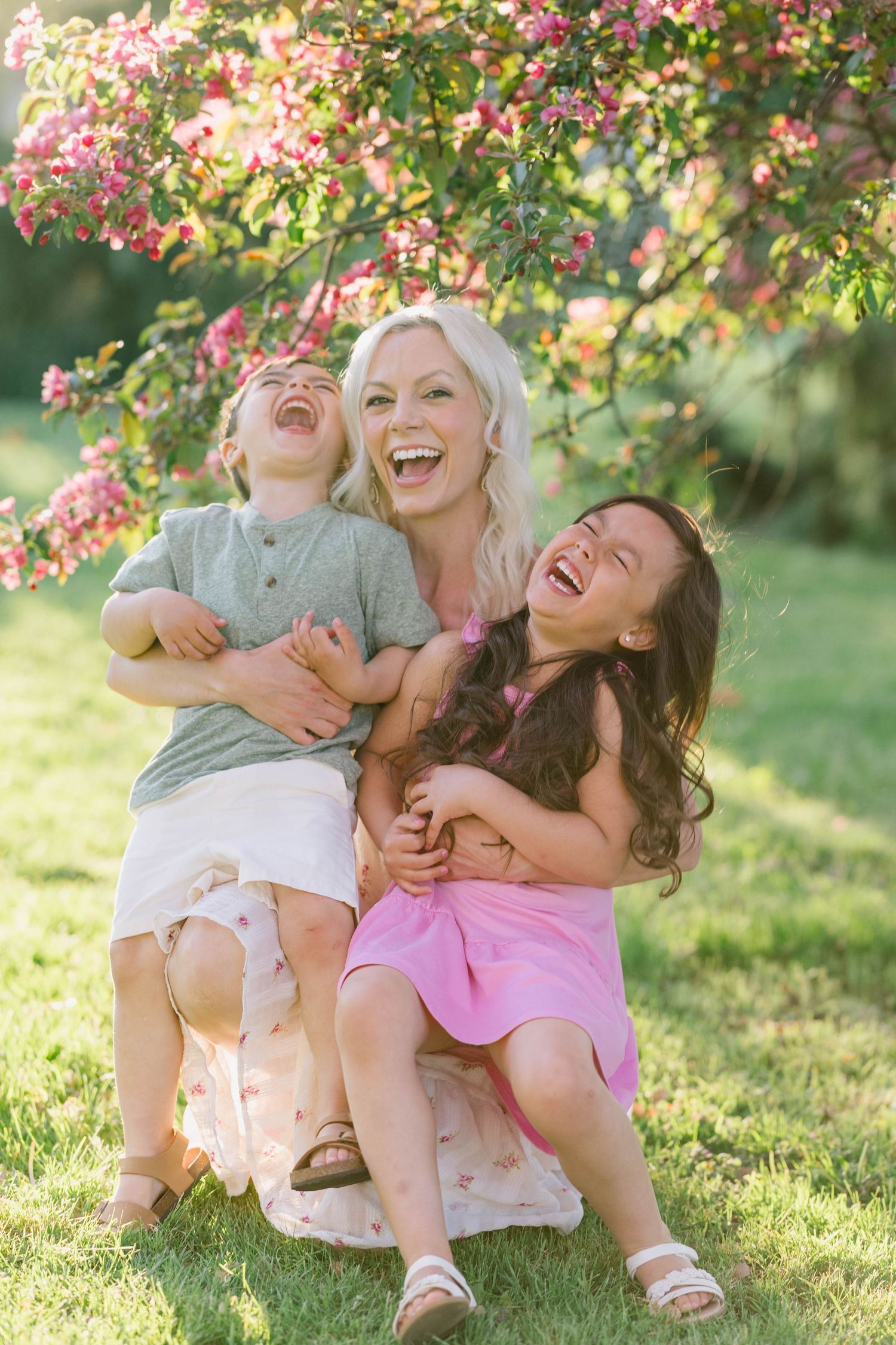 Blonde mother laughs joyfully with two children under pink flowering tree in sunny garden.