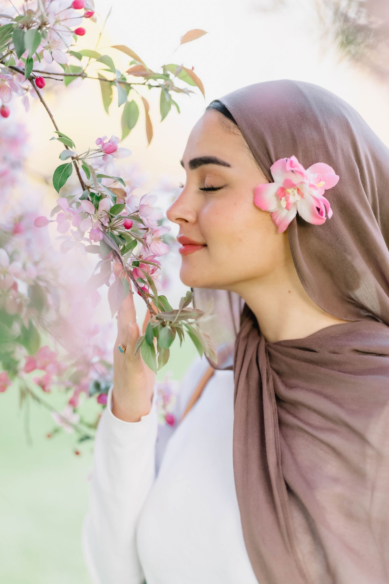 Woman in hijab smelling pink cherry blossoms with flower tucked behind ear in spring garden.