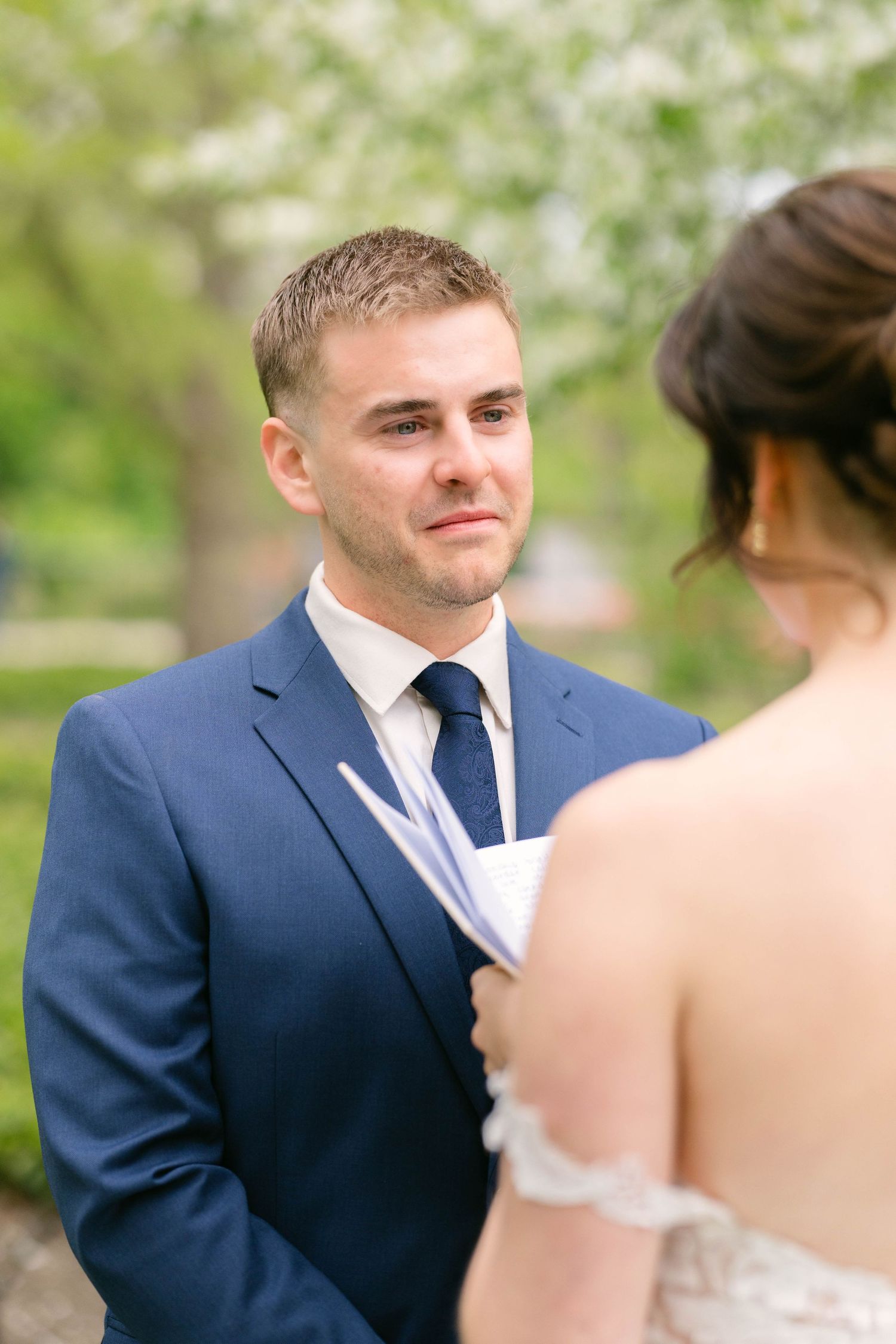 Emotional groom in navy suit tears up while listening to bride read wedding vows outdoors.