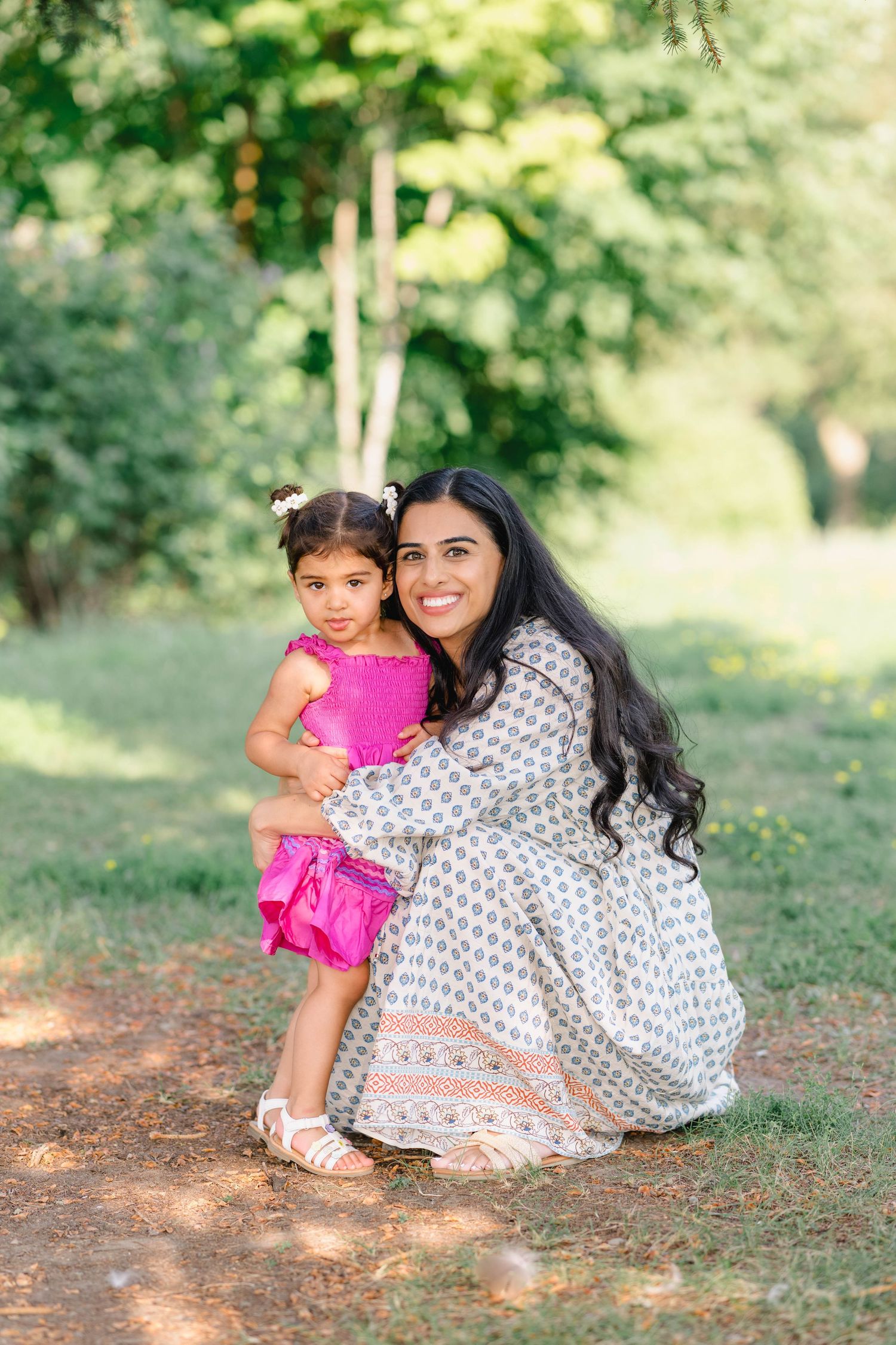 A smiling woman in a floral dress crouches to hug a young girl in pink outfit outdoors in a green park.