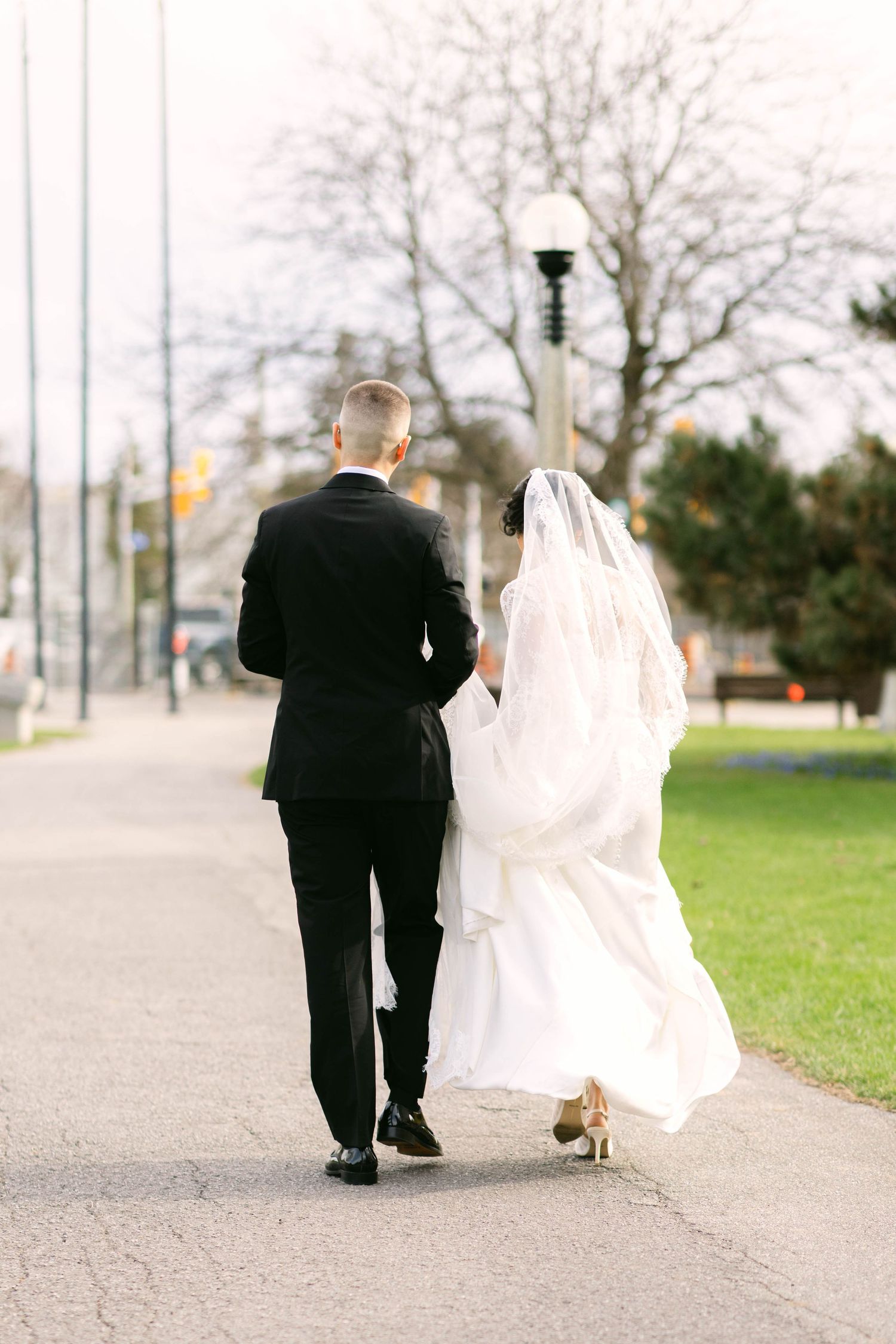 Bride and groom walking hand in hand down a path, viewed from behind, bride in white gown with veil, groom in black tuxedo.