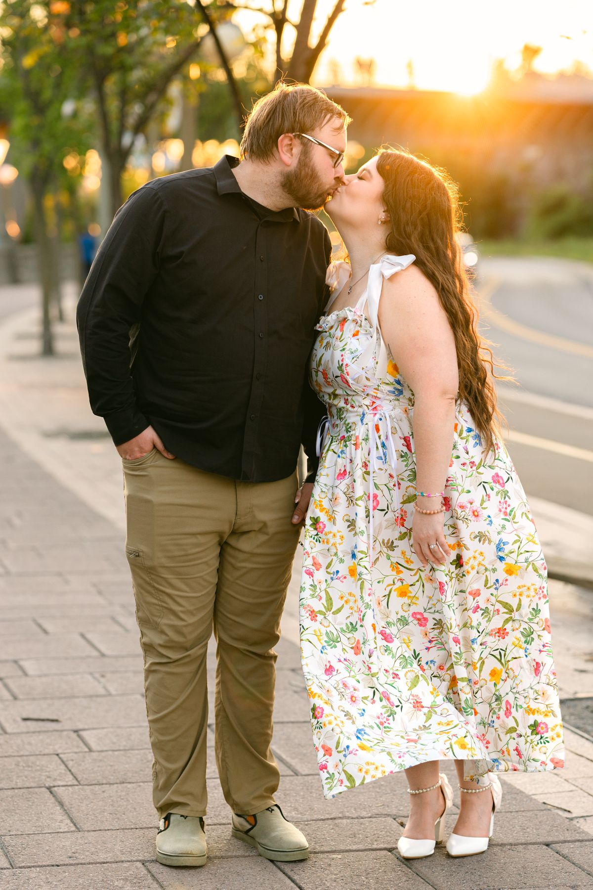 Couple sharing a romantic kiss at golden hour on a city sidewalk, woman in floral dress and man in black shirt.