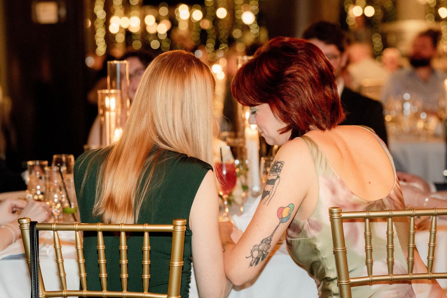 Two women with tattoos sit together at an elegant candlelit event, sharing a private conversation.