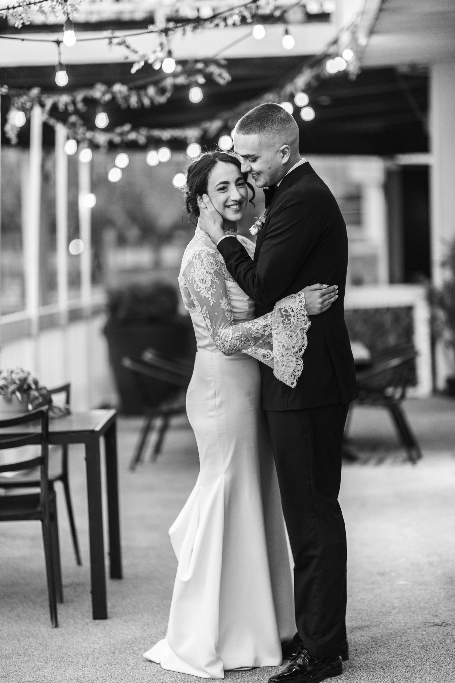 Black and white photo of smiling bride in lace gown embracing groom in suit at outdoor wedding reception.